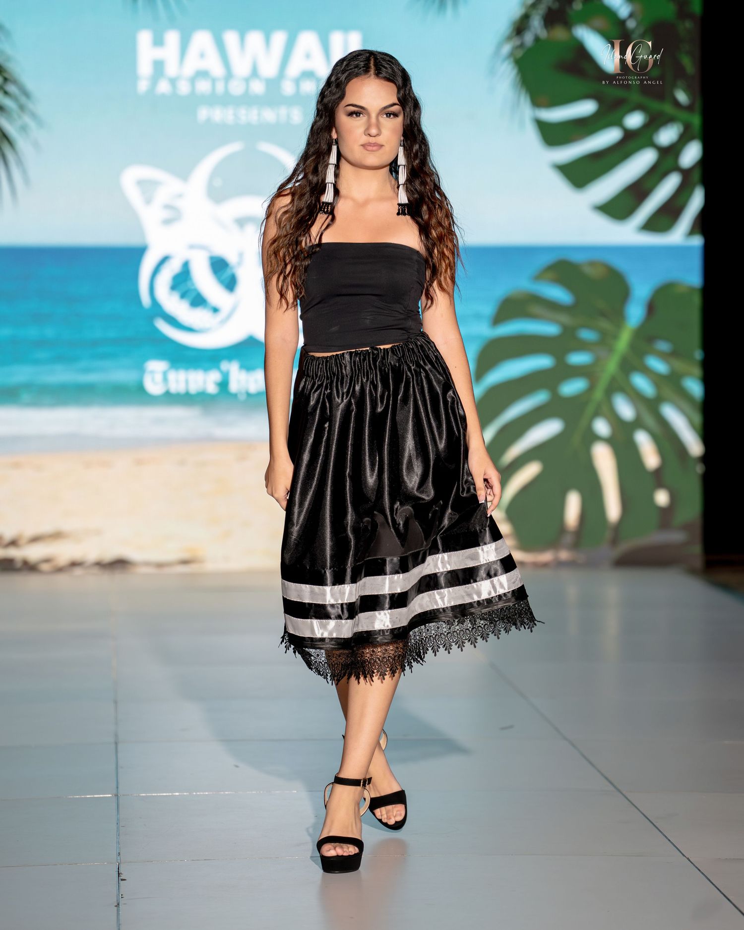 A model walks the runway in a black babydoll dress with white trim detail at a tropical-themed fashion show.