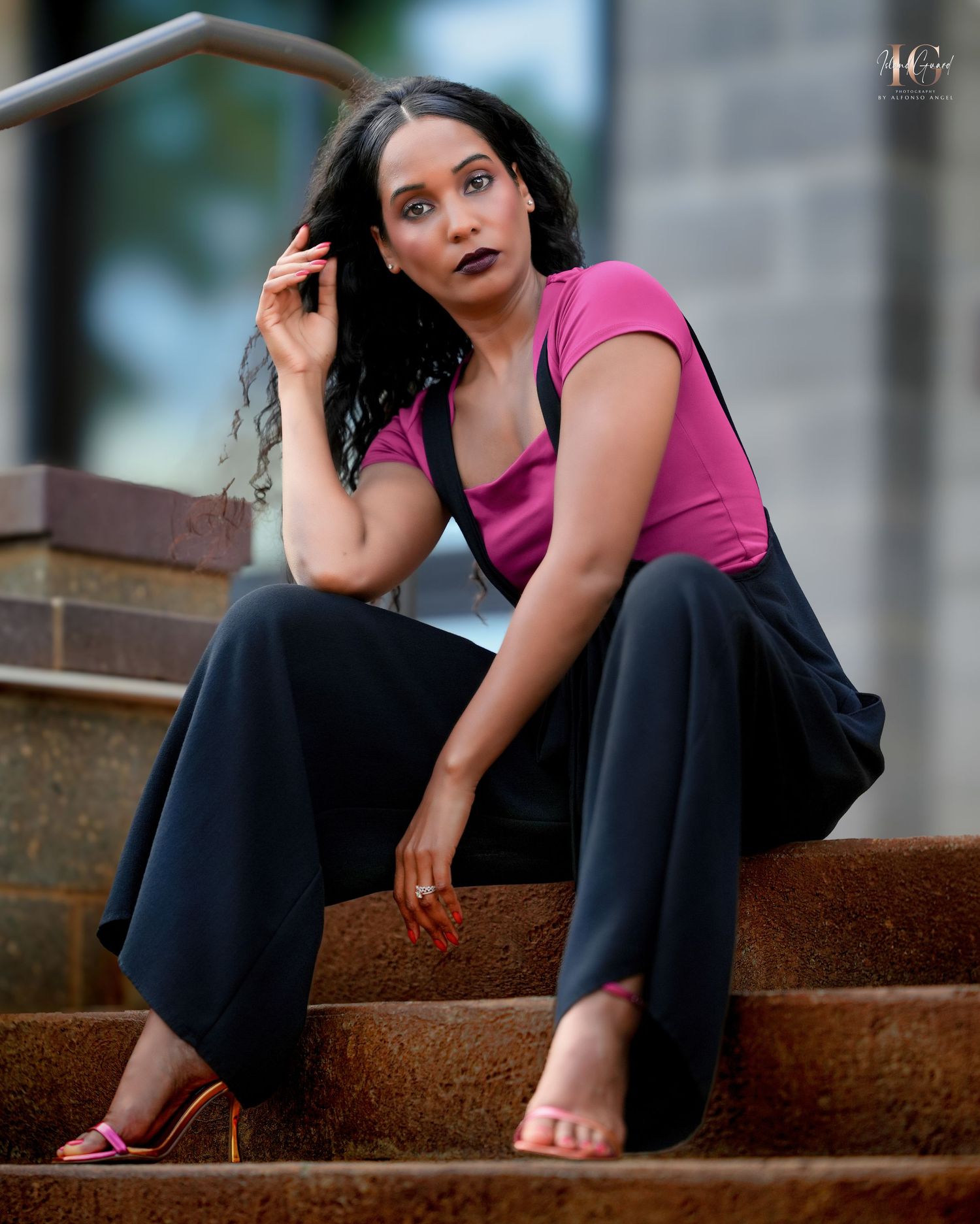 A model in a bright pink top and dark palazzo pants poses thoughtfully on concrete steps outdoors.