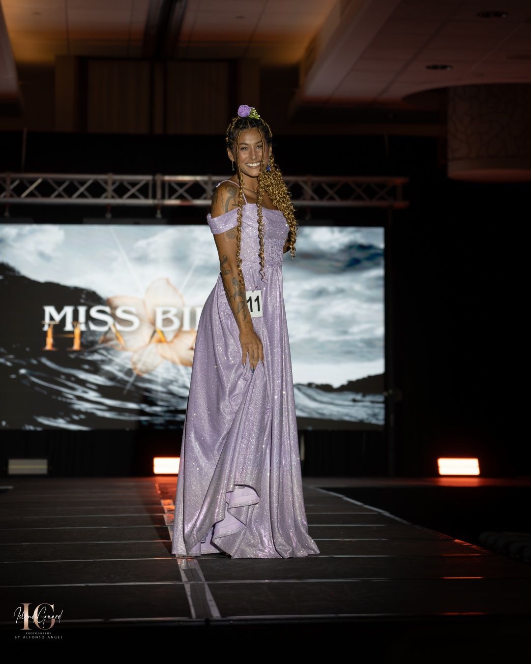 Model walks down runway in flowing lavender gown with dramatic cape during evening wear fashion show segment.