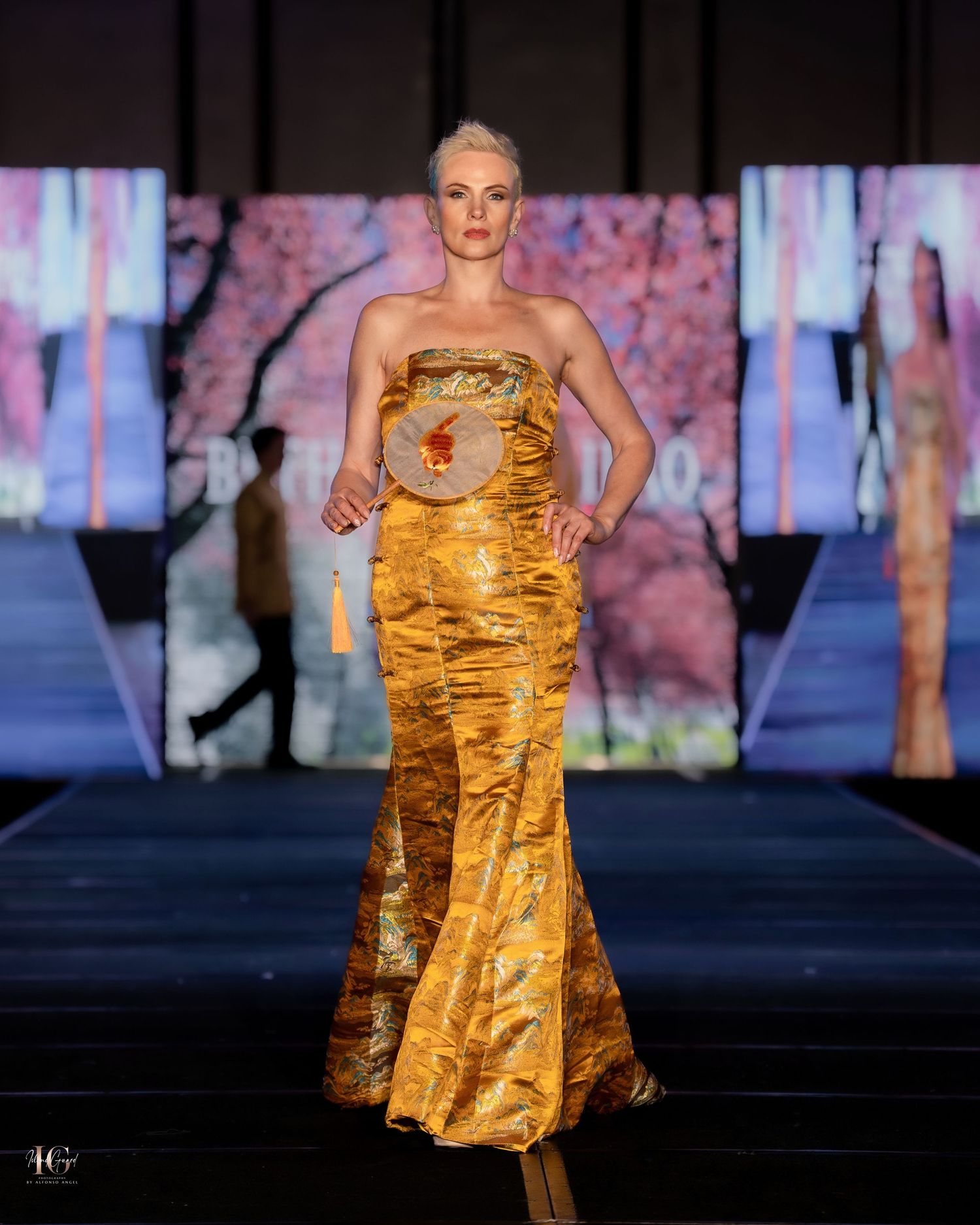 Model walks runway in strapless gold metallic mermaid gown against vibrant digital backdrop during fashion show.