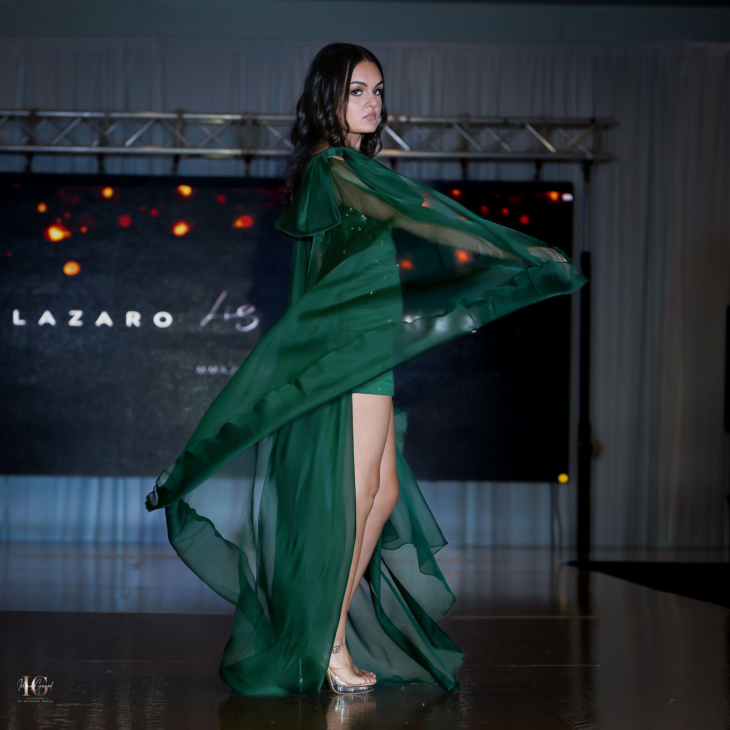 Model twirls in flowing emerald green evening gown with dramatic cape detail.
