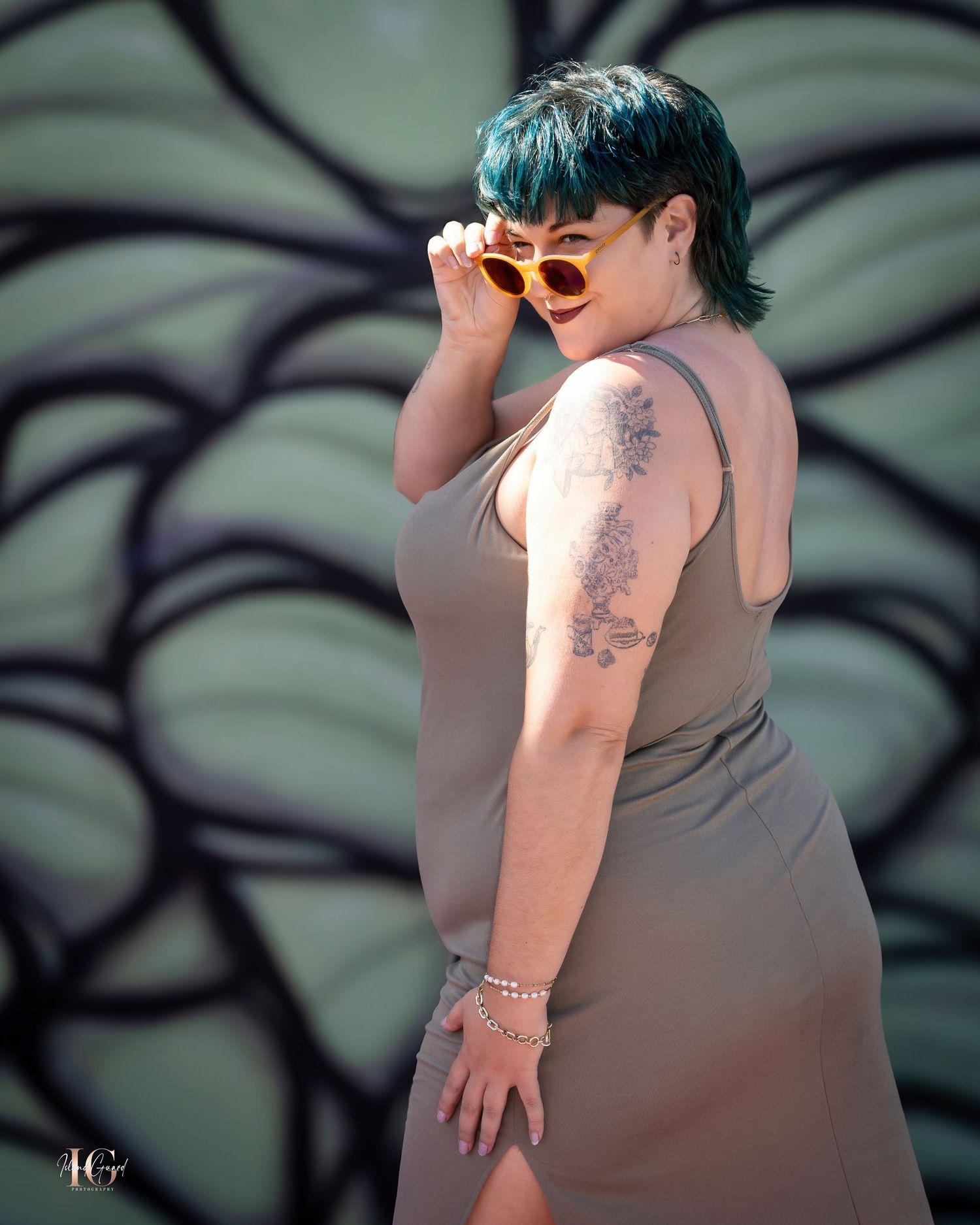Person with green hair and sunglasses poses in beige dress against decorative wall background.