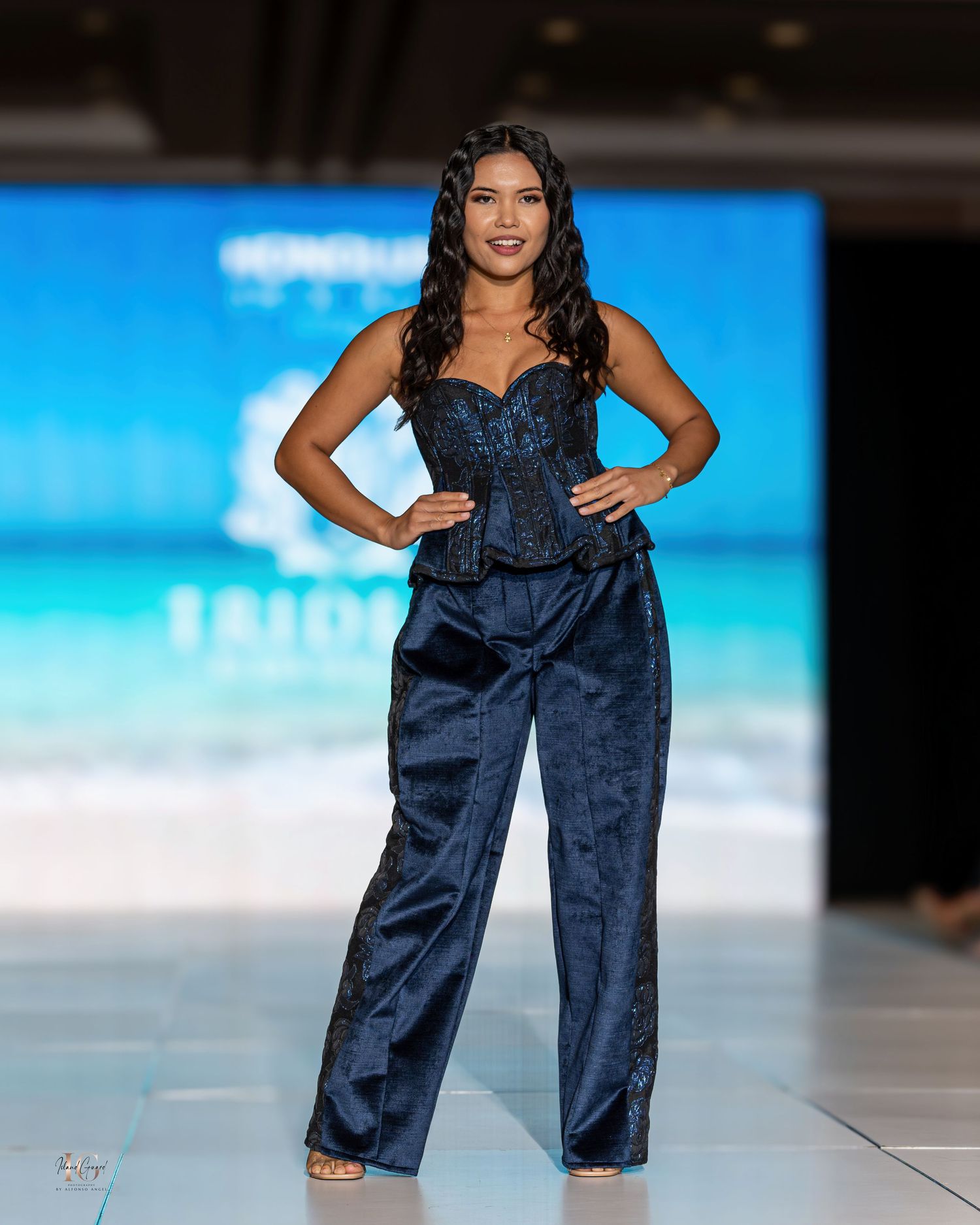 Model showcases navy blue jumpsuit with ruffled neckline on tropical-themed runway.
