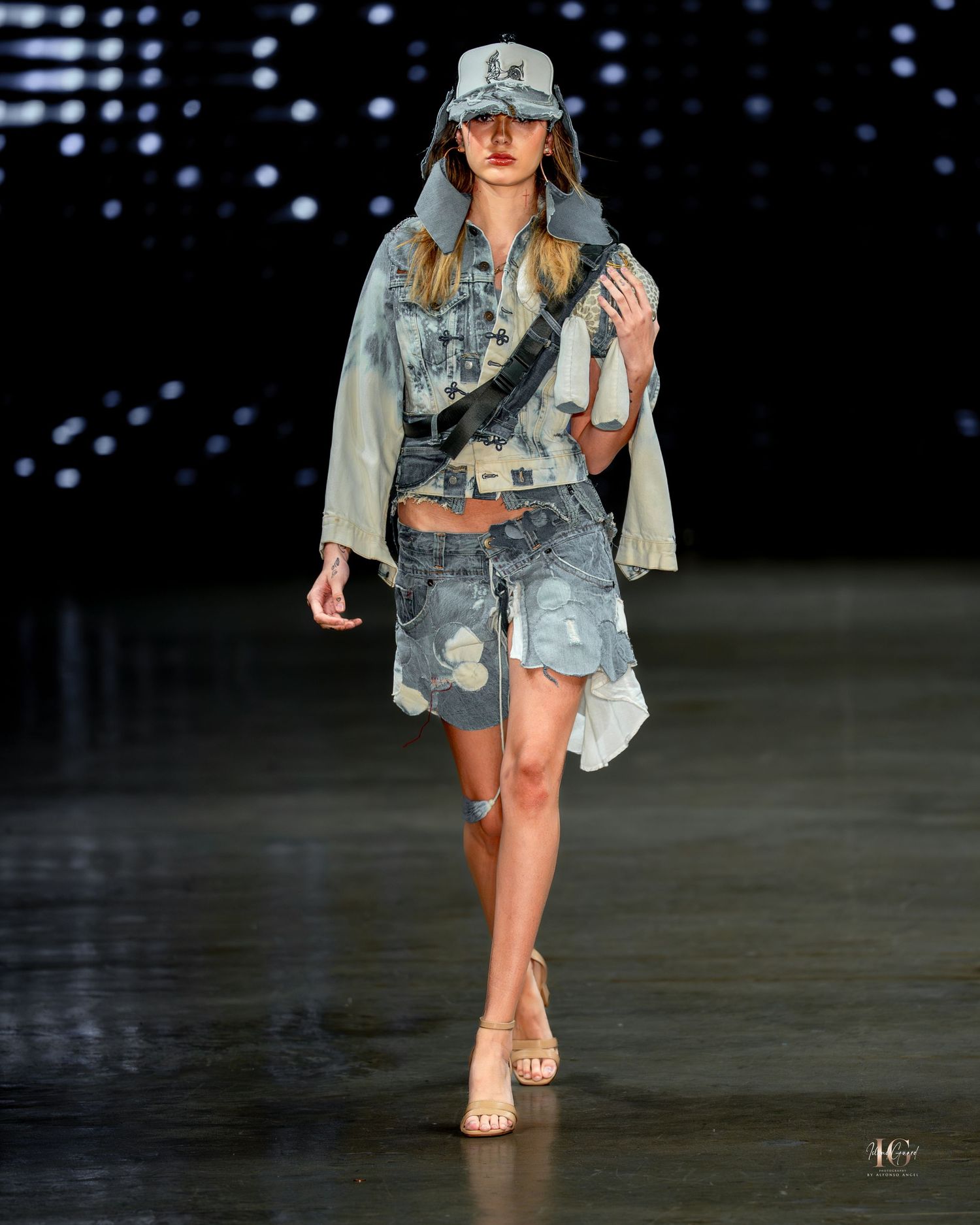 Model walks runway in distressed denim jacket and shorts with baseball cap during fashion show.