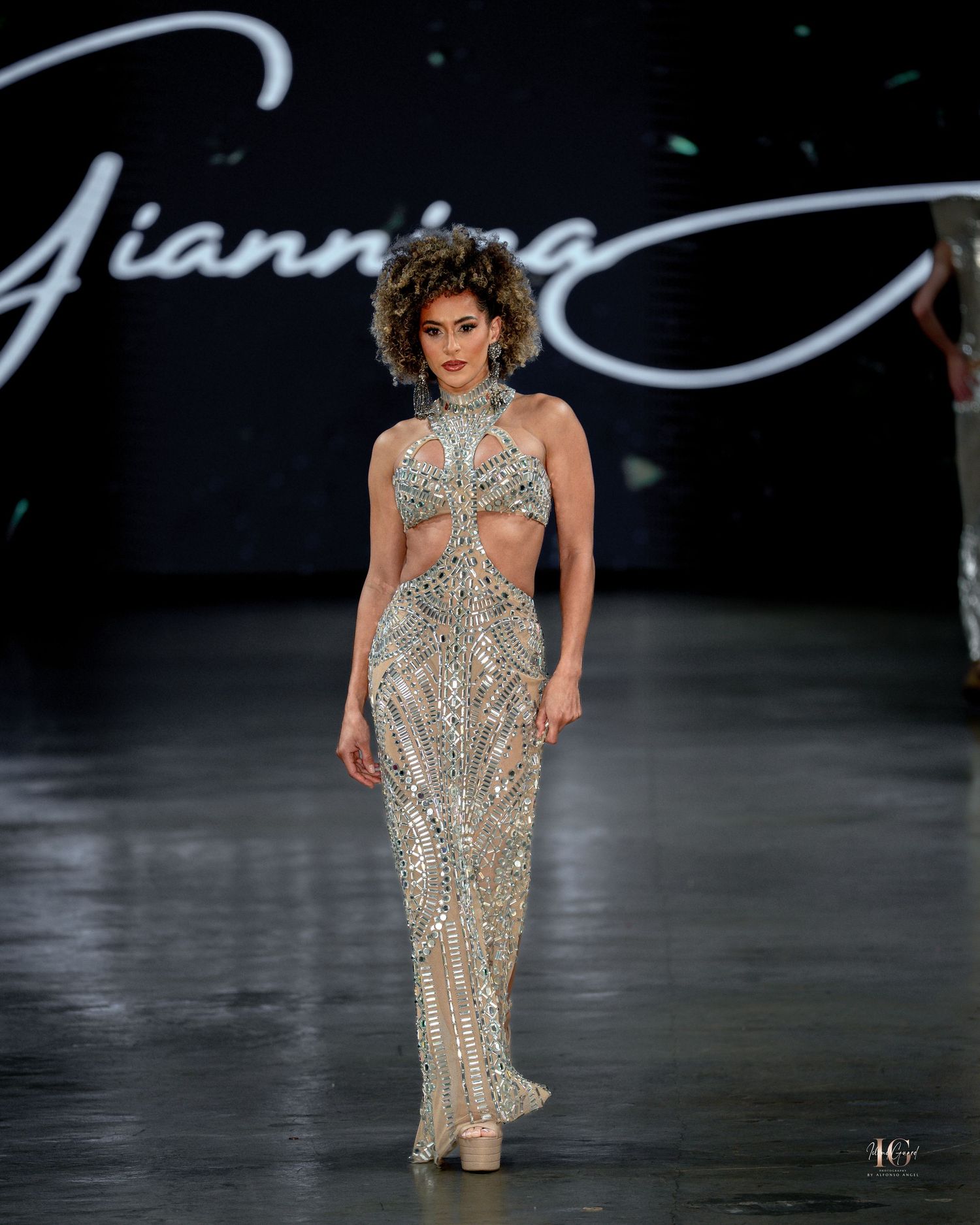 Model walks runway in shimmering silver evening gown with halter neckline and crystal embellishments during fashion show.