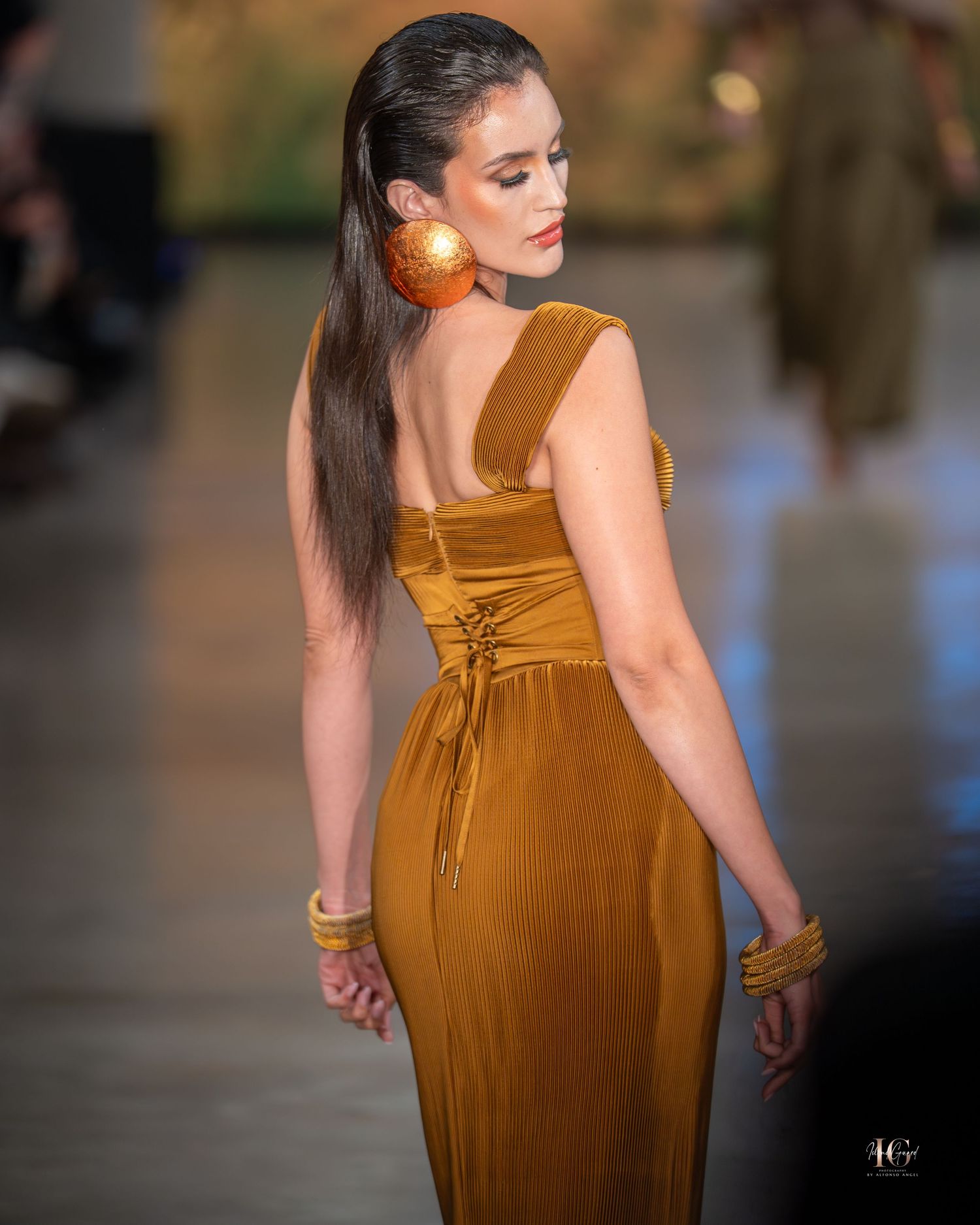 Model wearing a form-fitting mustard yellow dress with lace-up back detail and statement orange earrings on the runway.