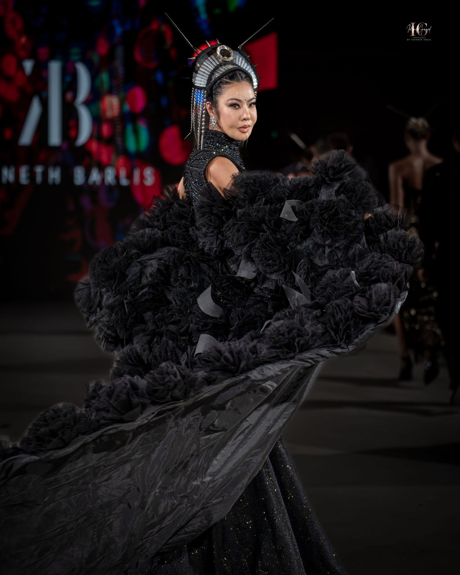 A model walks the runway in a dramatic black ruffled gown with flowing train at a fashion show.