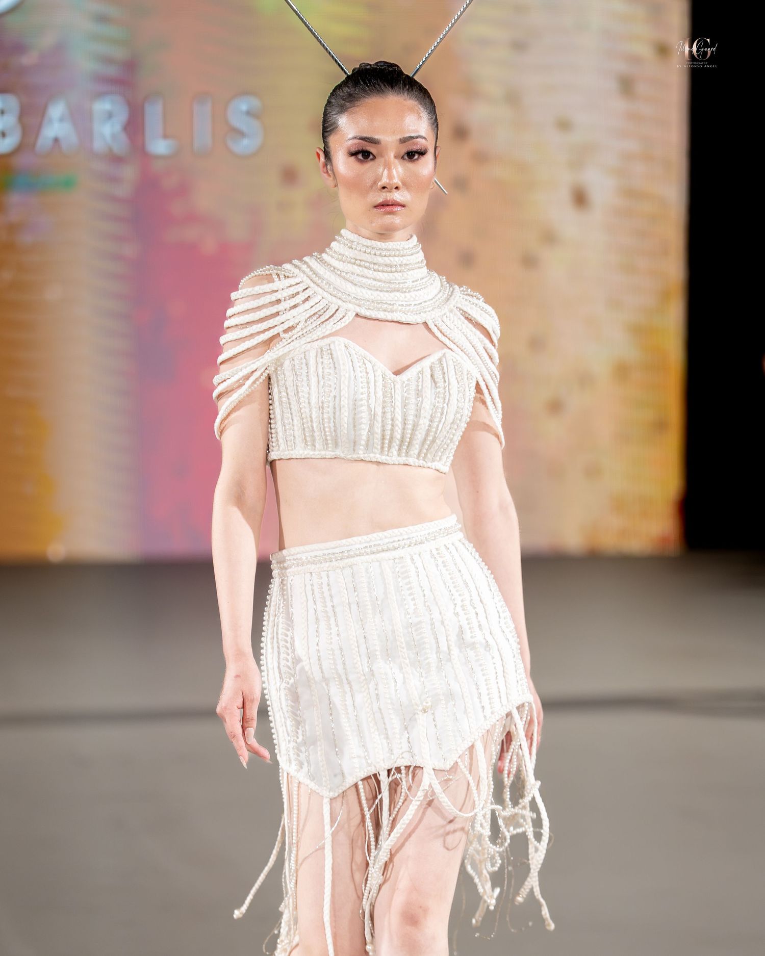 Model walks runway in white two-piece outfit with high neck crop top and fringed skirt at fashion show.