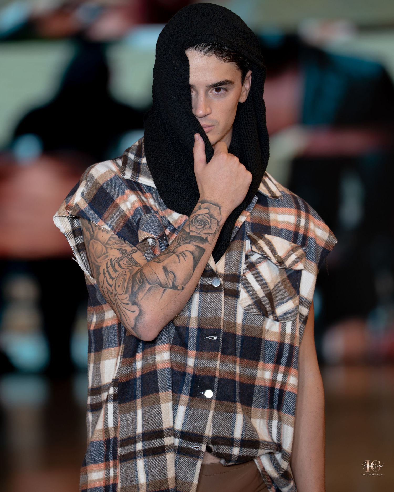 Model wearing plaid sleeveless shirt with tattoos poses against blurred background in fashion photoshoot series.