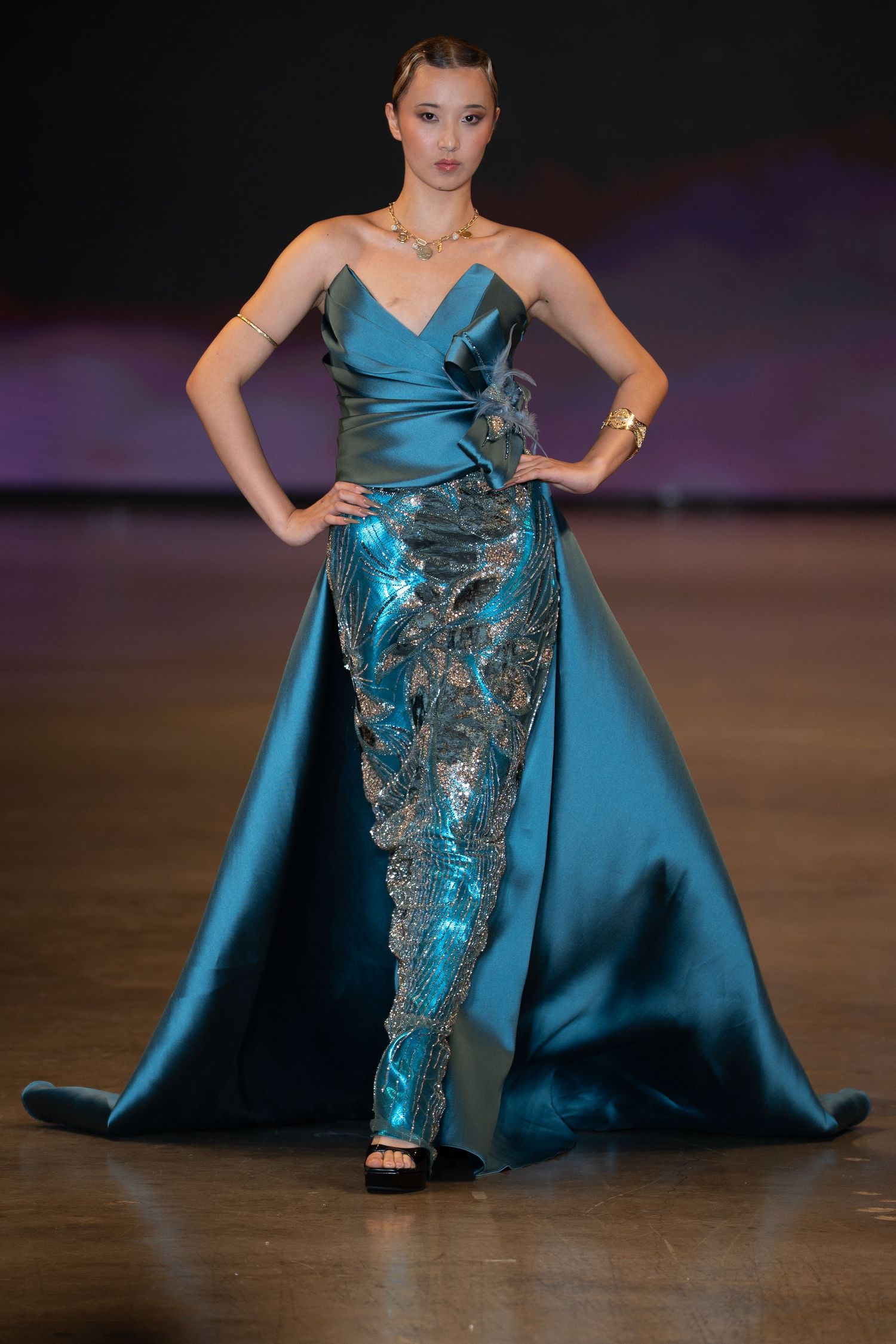 Model showcases elegant teal evening gown with metallic embellishments on dark runway stage.