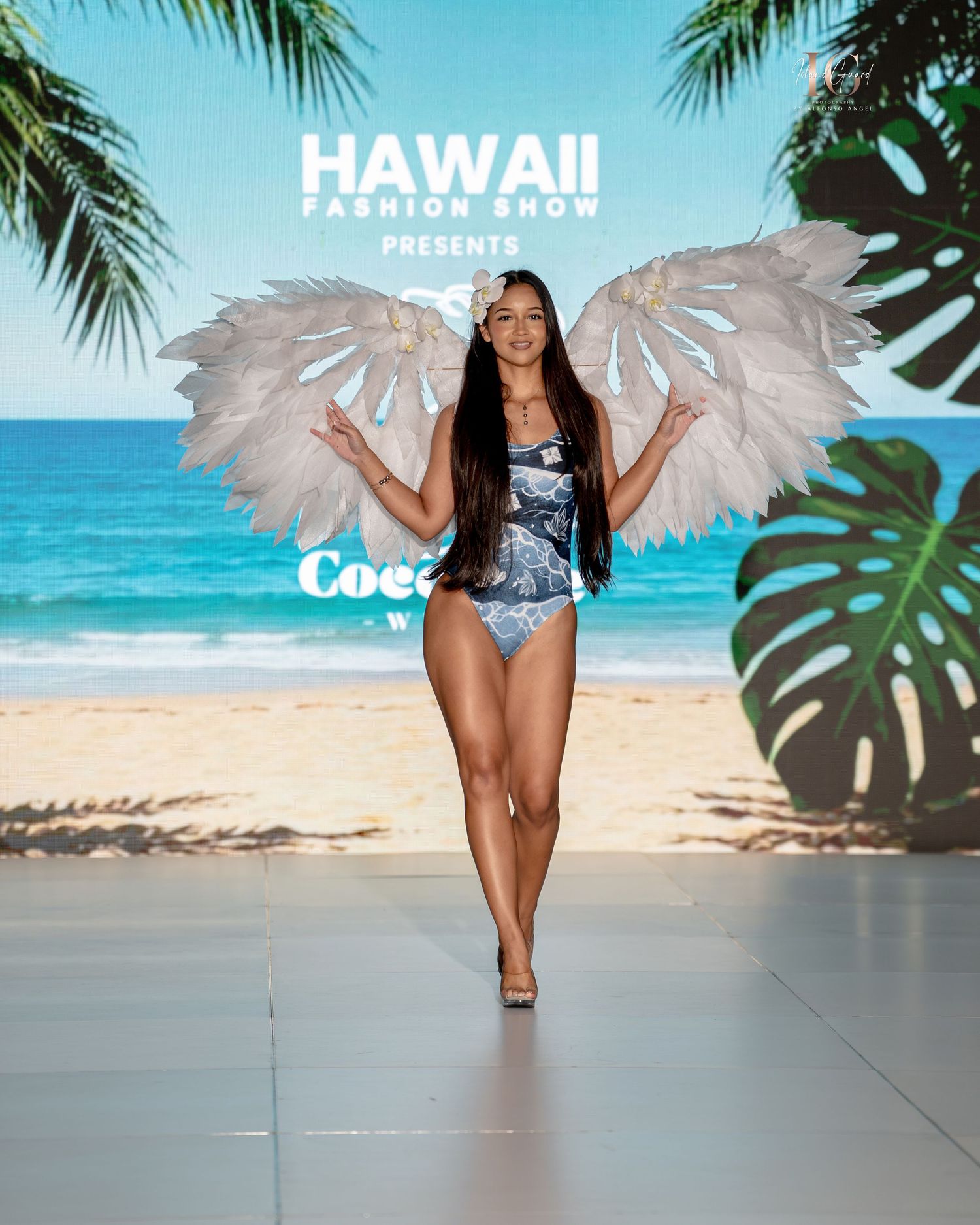 A model walks the runway in a blue swimsuit at a Hawaii fashion show with tropical beach backdrop and palm fronds.
