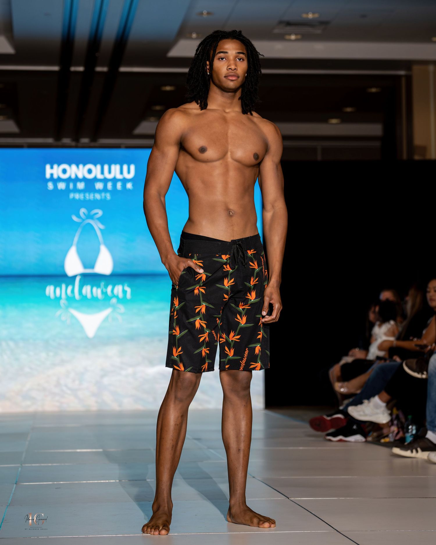 Male model showcases black and orange floral swim shorts on runway.