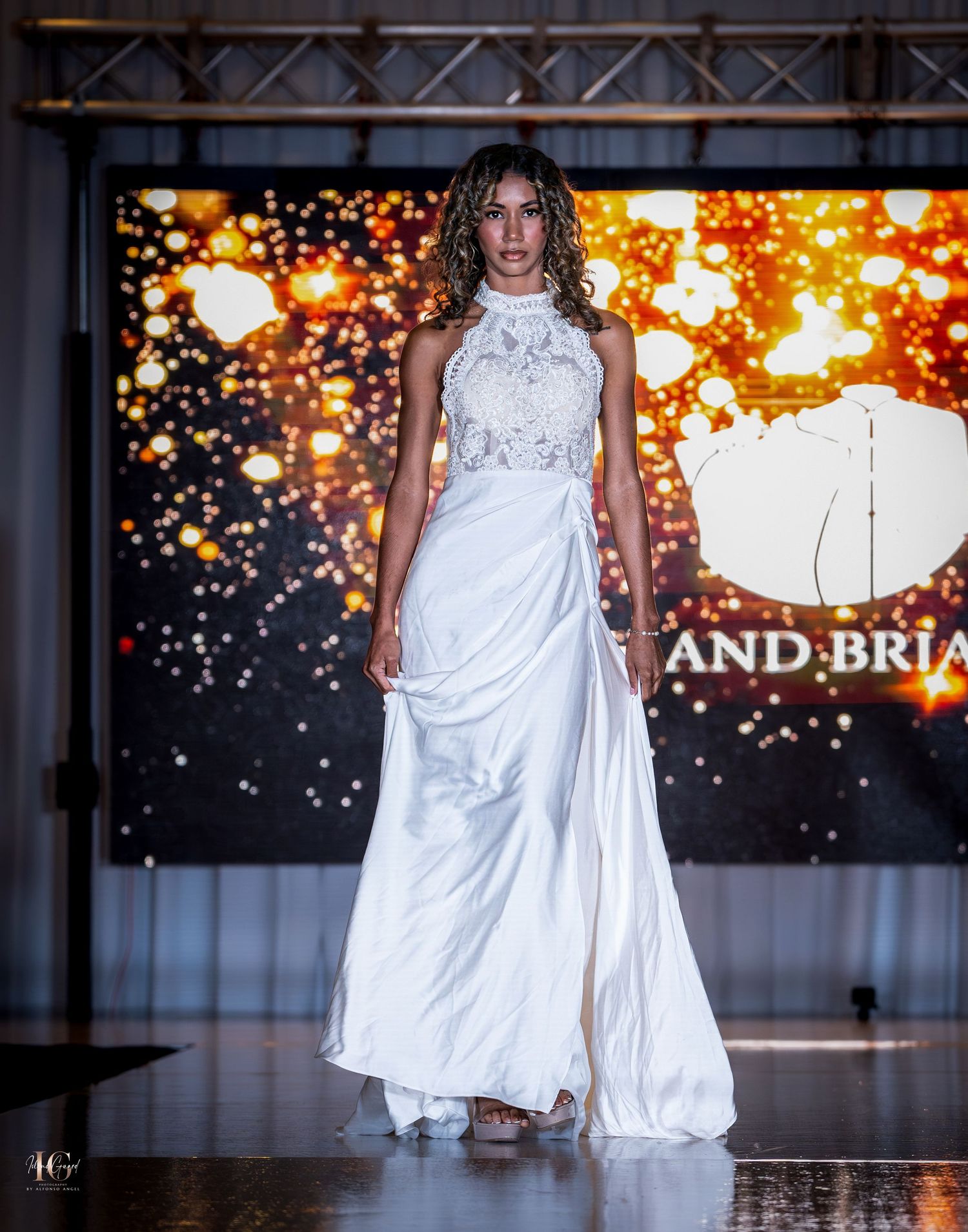 Models showcase flowing white formal gowns against sparkly backdrop during evening fashion show.