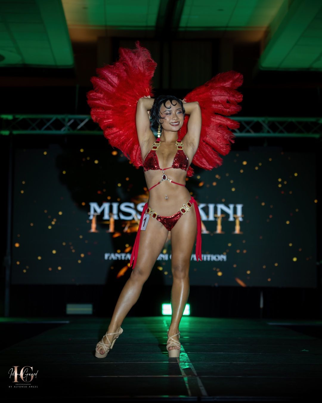 A performer in a red feathered headdress and bikini strikes a pose on a runway stage with spotlights and signage.