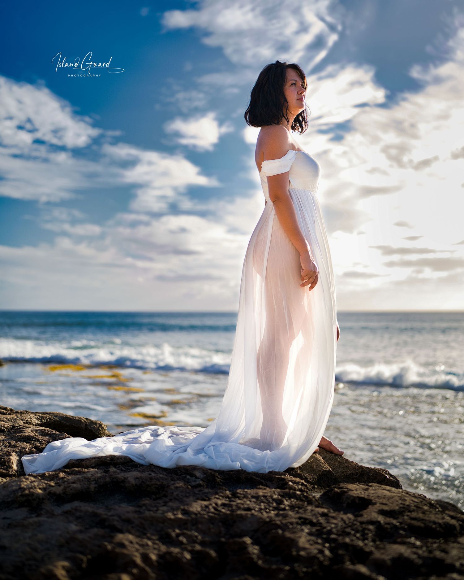 A figure in a flowing white dress stands on coastal rocks at sunset as ocean waves crash against the shore.