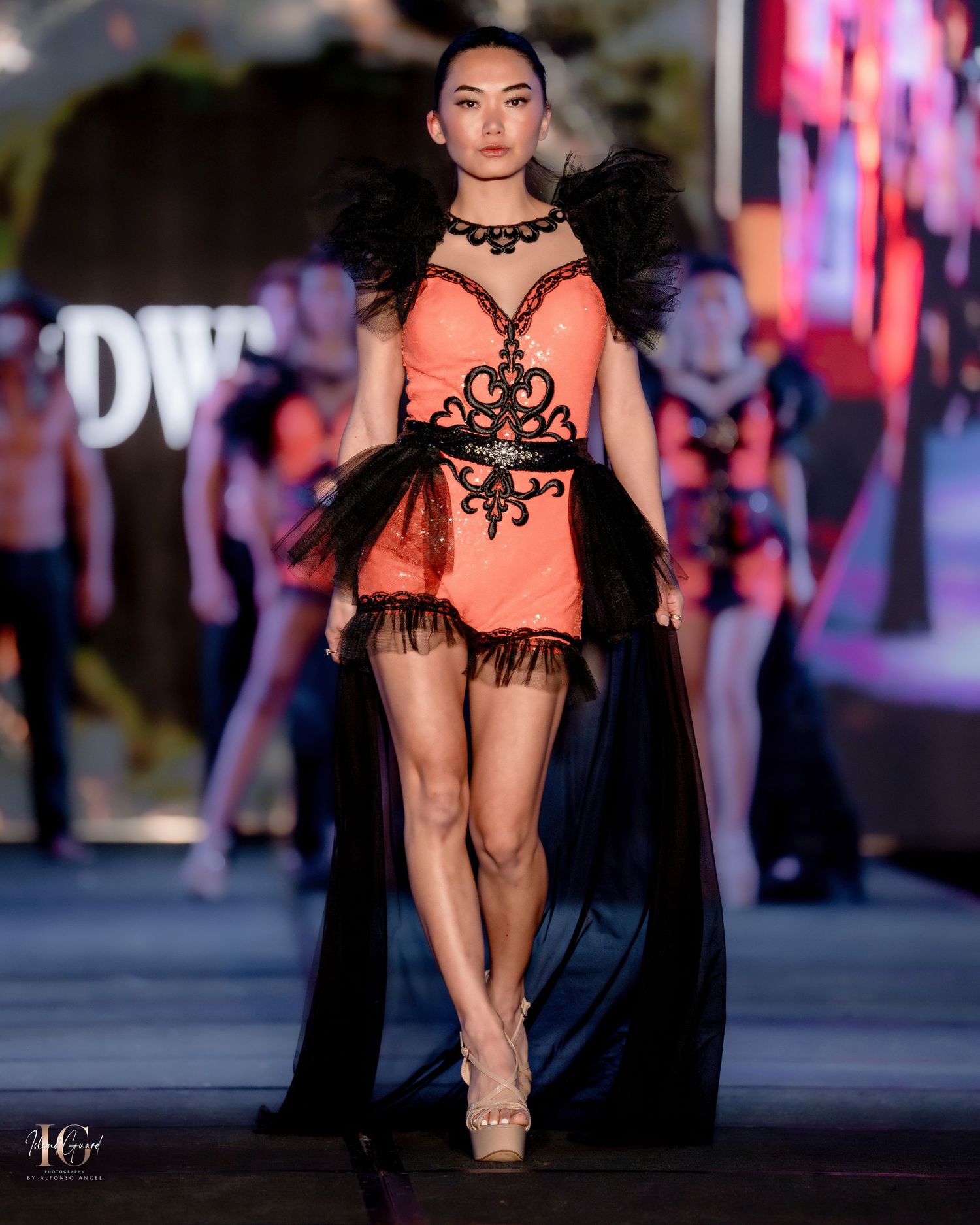 Fashion model walks runway in coral gown with black lace details and flowing cape.