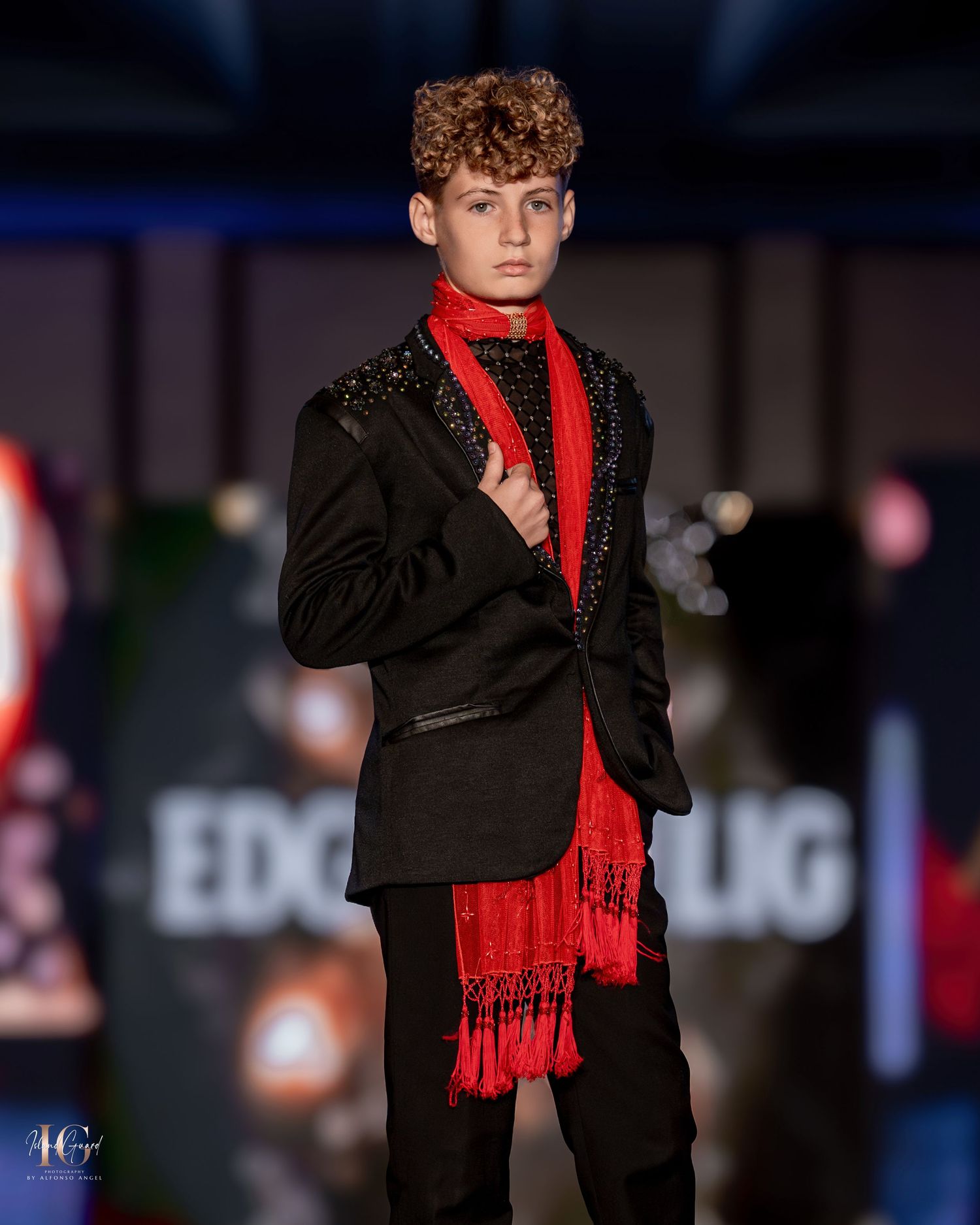 Fashion model wearing black suit with bright red scarf on dark runway stage.