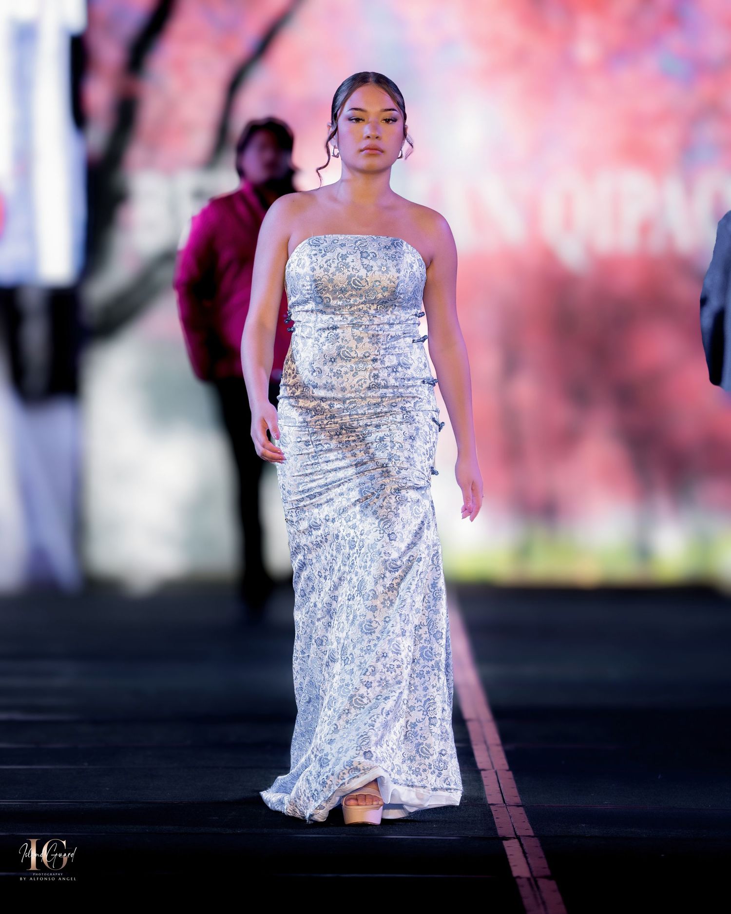 Model walks runway in strapless white floral evening gown with fitted silhouette against pink backdrop.