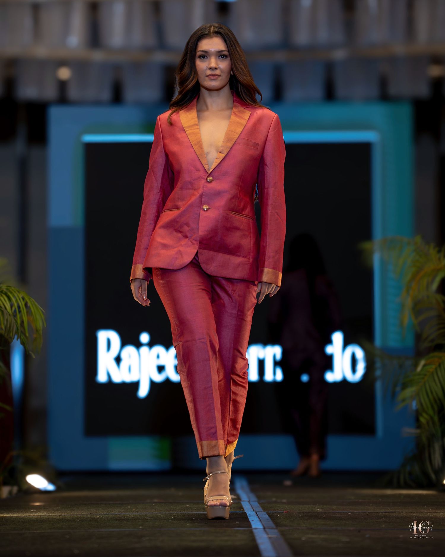 Model walks runway wearing coral-colored sheer blazer and matching pants during fashion show presentation.