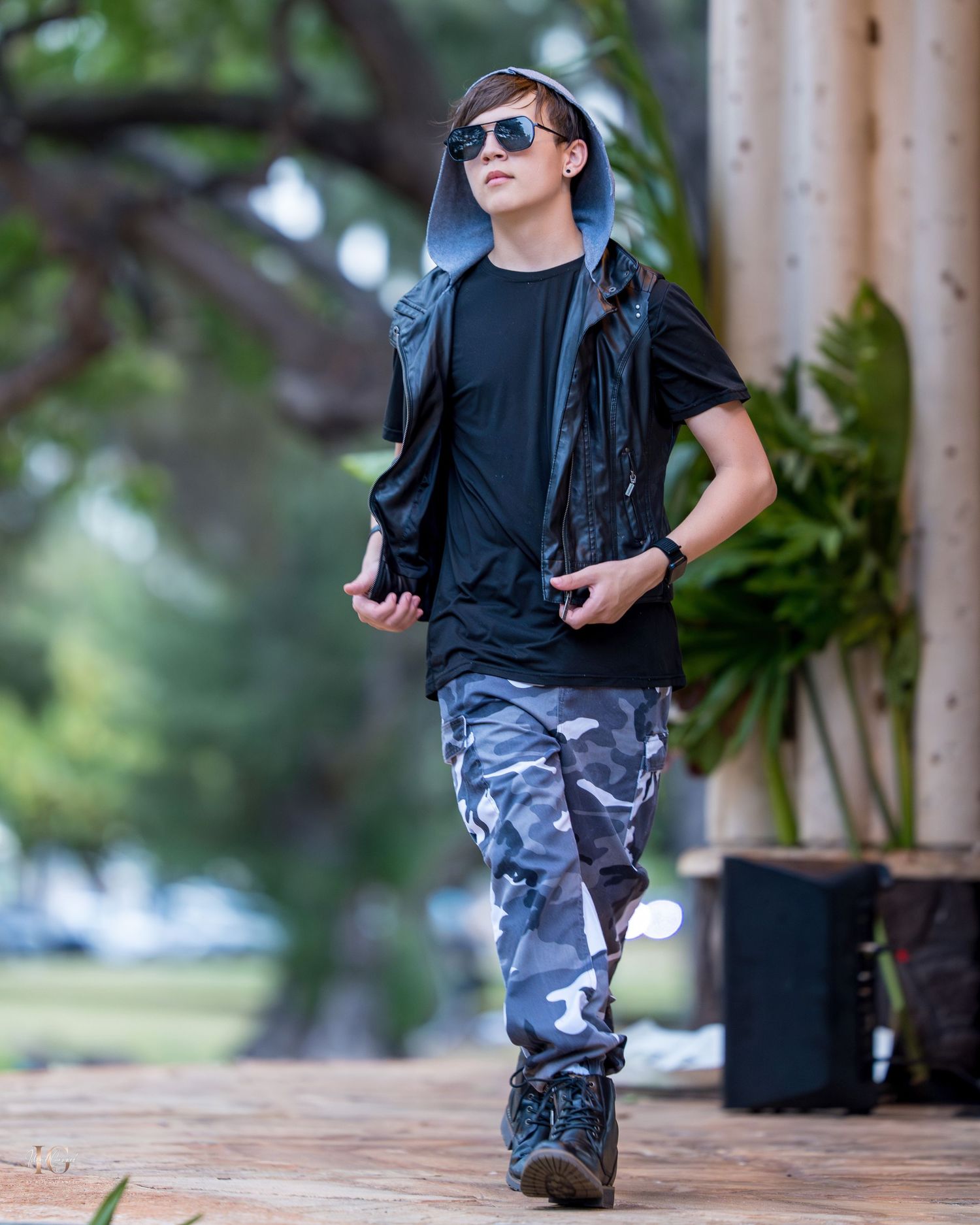 A young child in camo pants and sunglasses walks confidently on a wooden deck in an outdoor setting.