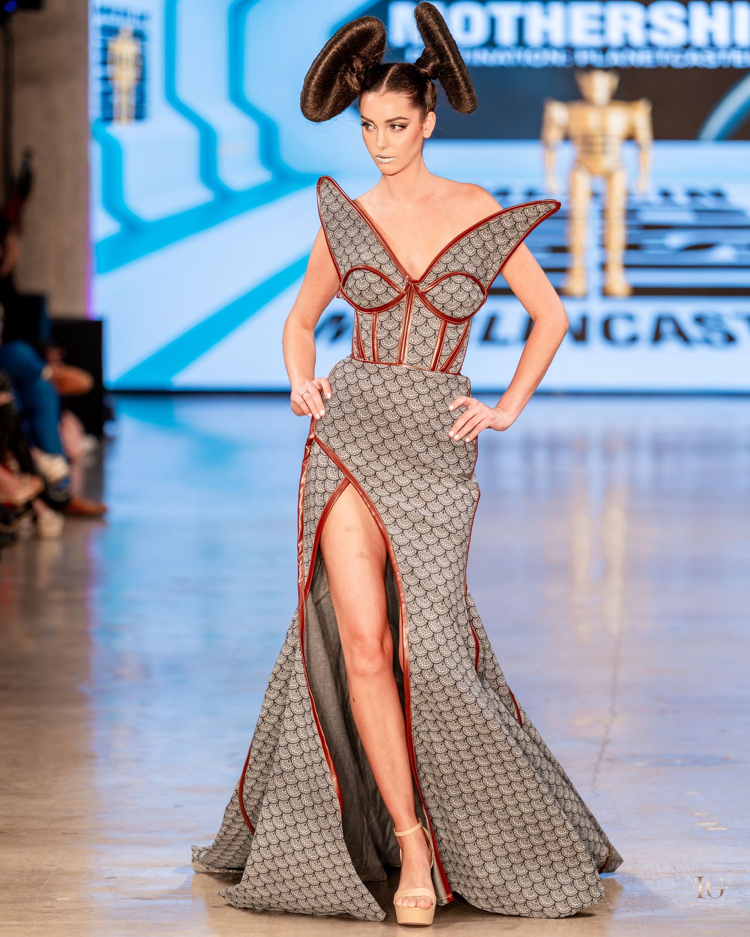 Model walks runway in gray patterned evening gown with high slit and dramatic neckline during fashion show.