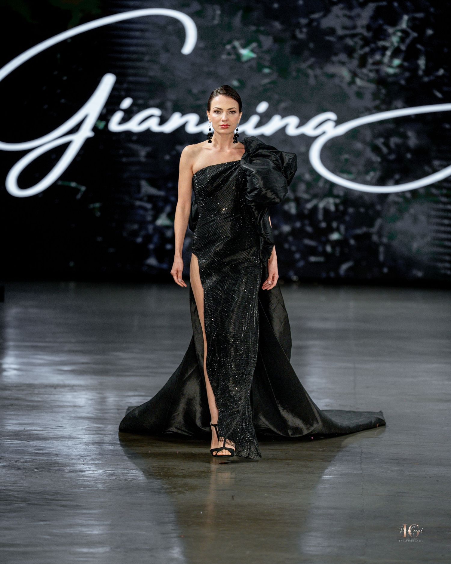 A dramatic black evening gown with flowing fabric sweeps across a glossy runway stage during a fashion show.