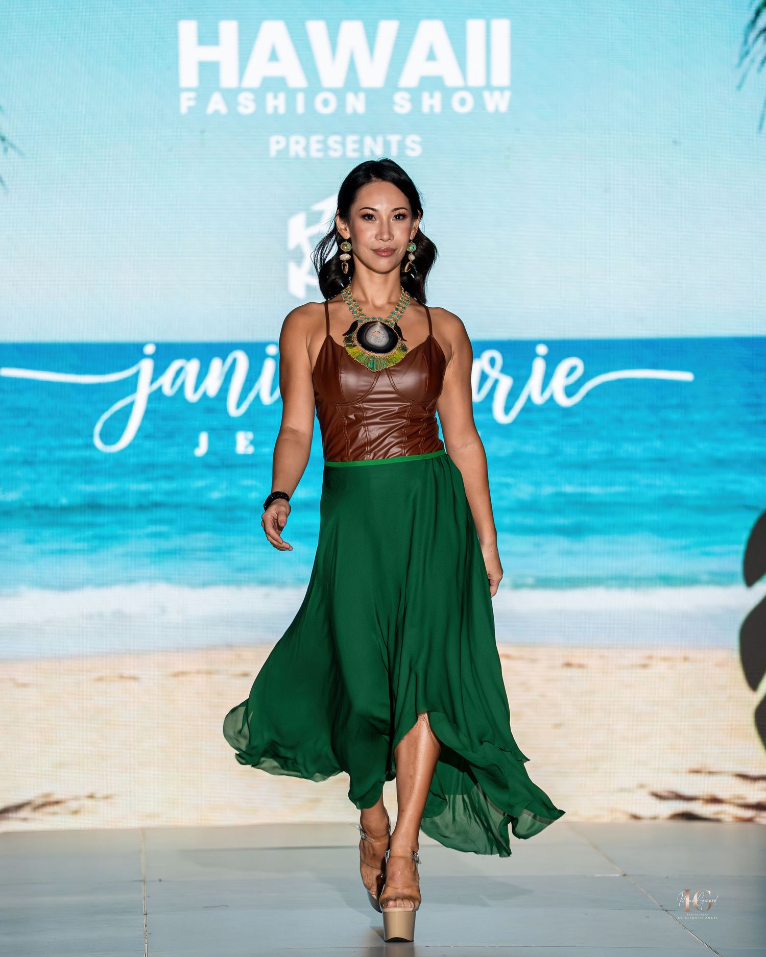 Model walks runway in brown leather top and flowing emerald green skirt at Hawaii Fashion Show against beach backdrop.