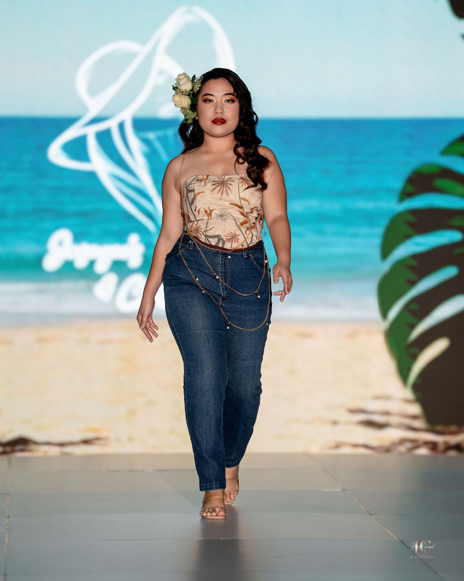 Model walks runway in beige floral top and high-waisted jeans at tropical-themed beach fashion show.