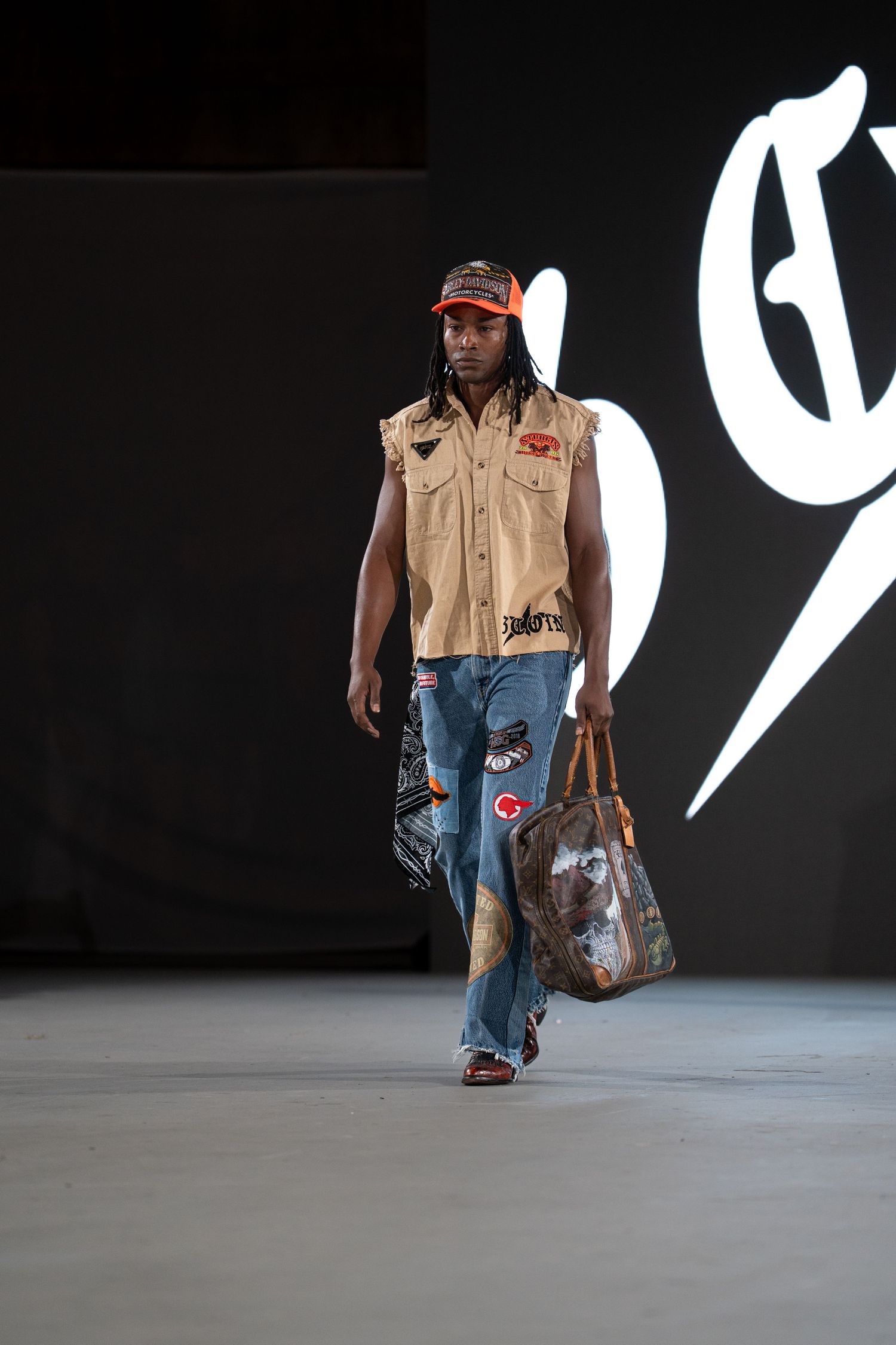 Fashion model walks runway in distressed denim jeans and tan vest during contemporary streetwear show.