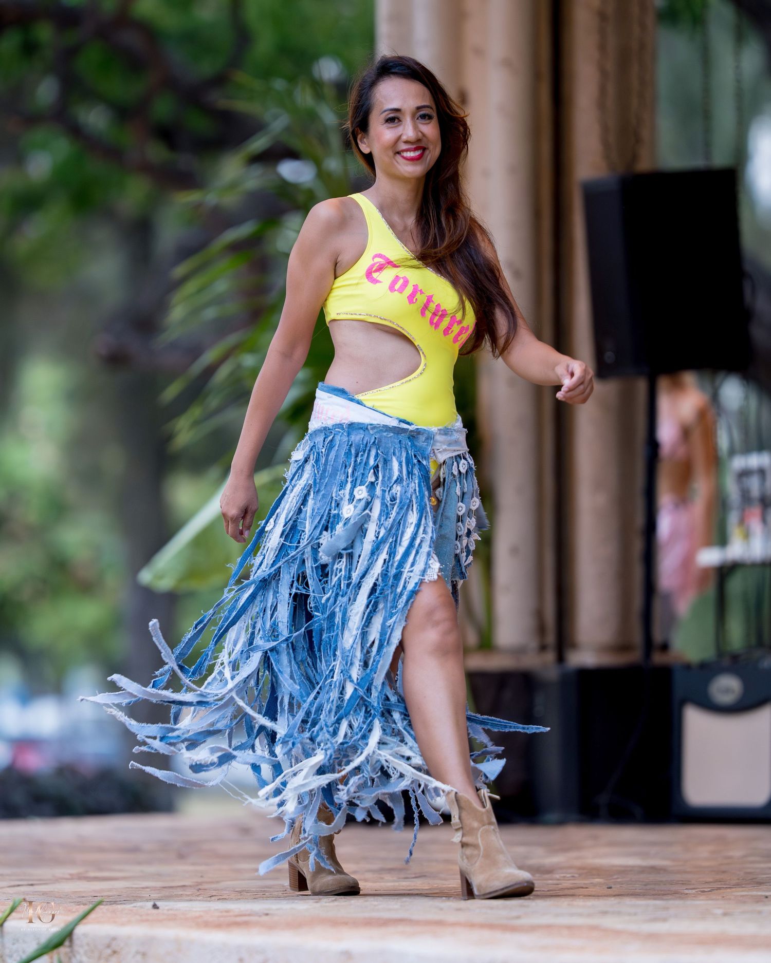 A dancer in a yellow top and blue fringed skirt performs dynamic movements on an outdoor stage.