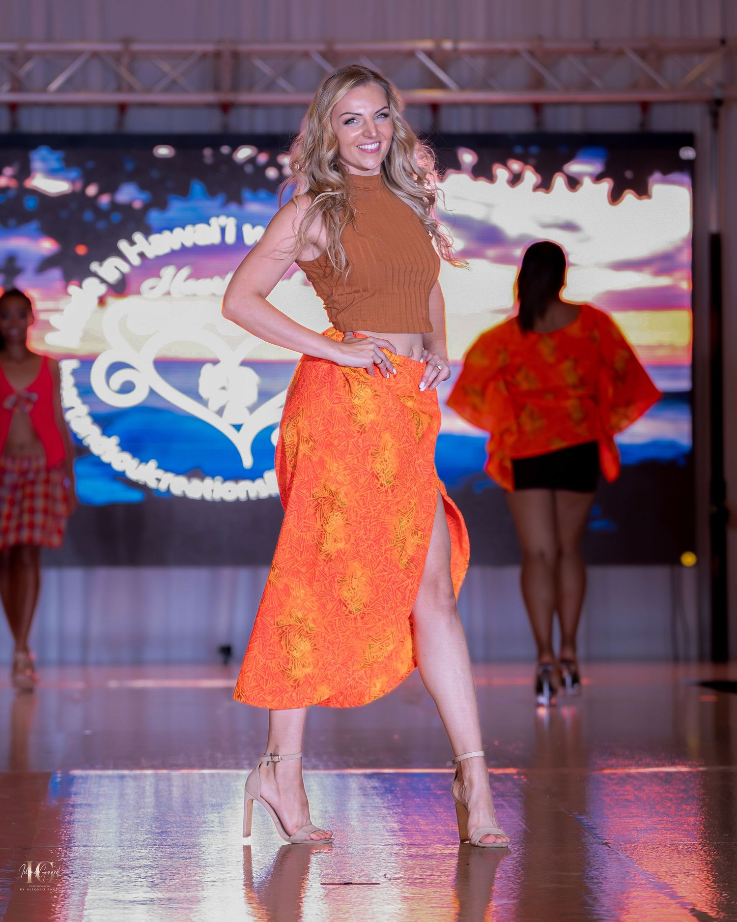 Models present bright orange summer dresses on an illuminated fashion runway.