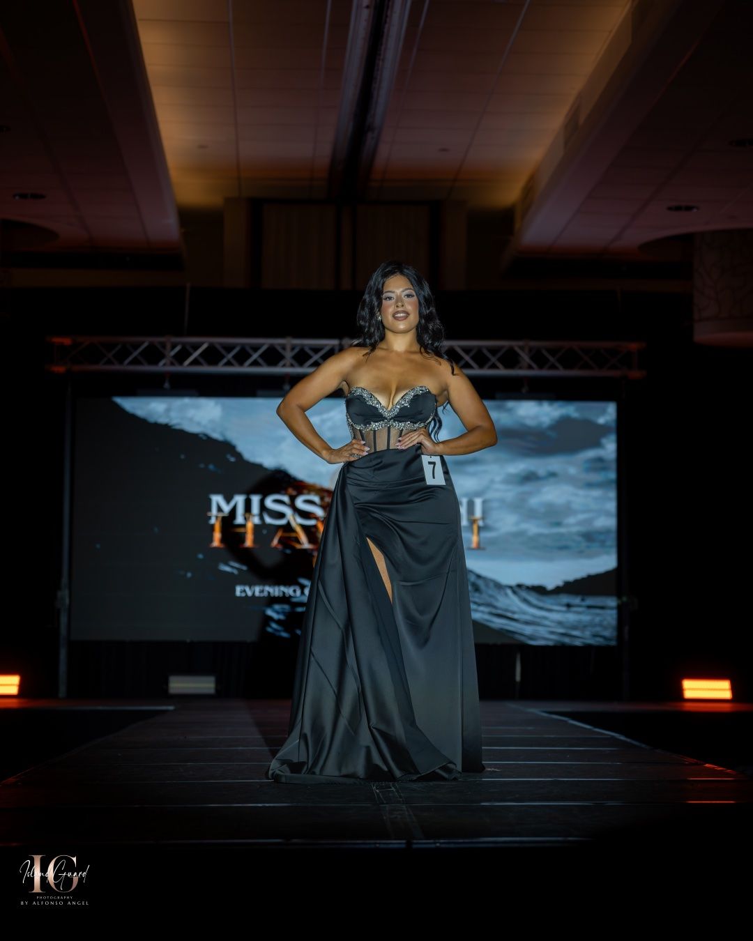 A contestant stands confidently on a fashion show runway in a shimmering black evening gown with stage lighting and decor.