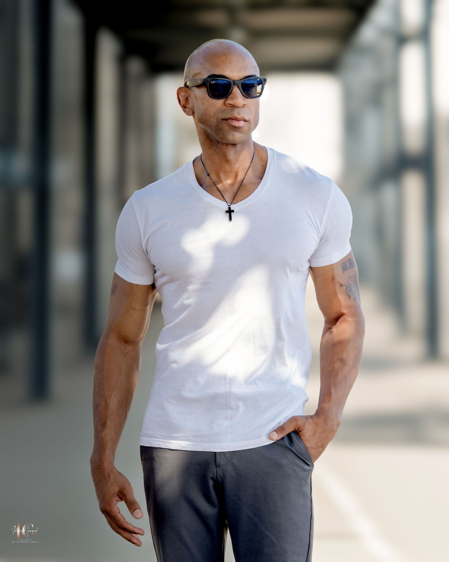 Muscular person in a white v-neck t-shirt and sunglasses poses confidently against a blurred architectural background.