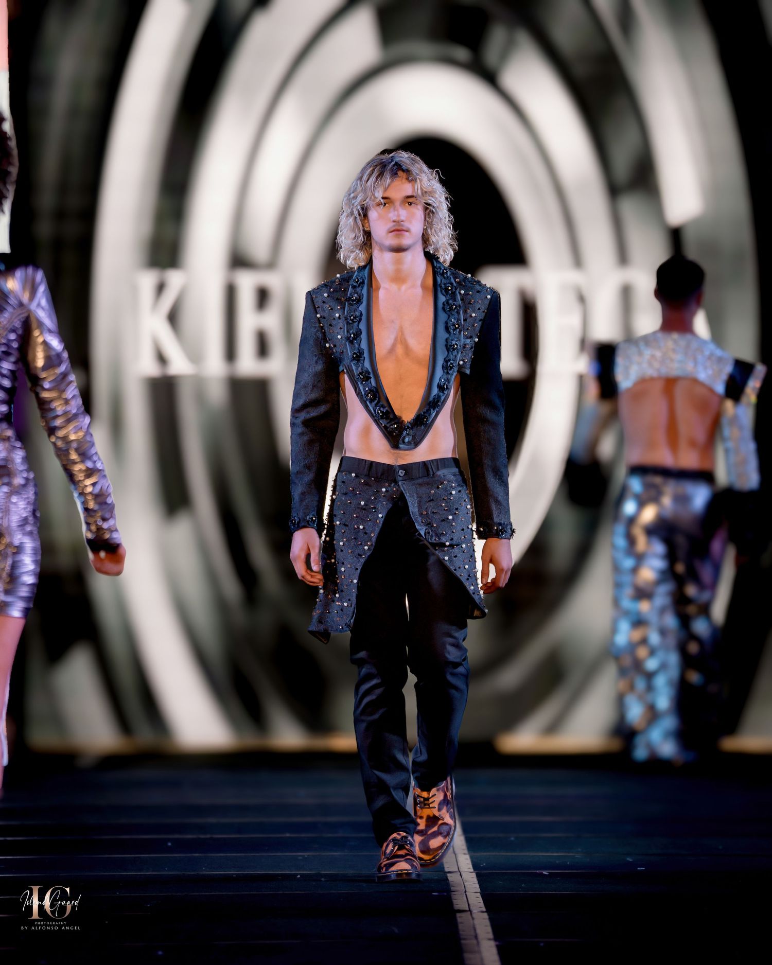 Model presents sparkly dark blazer with plunging neckline on illuminated runway.