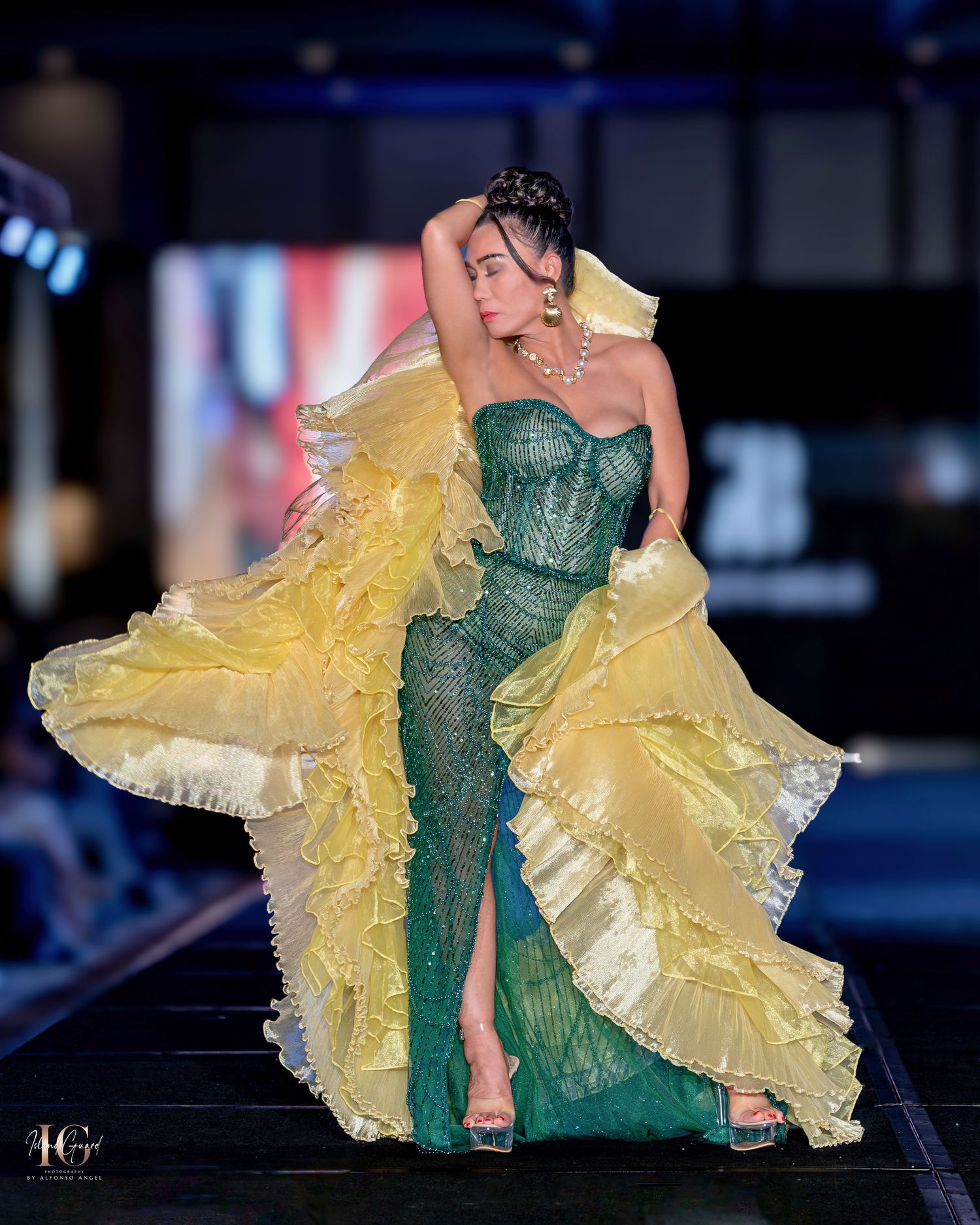 Emerald green fitted gown with yellow ruffled overlay displayed on runway.