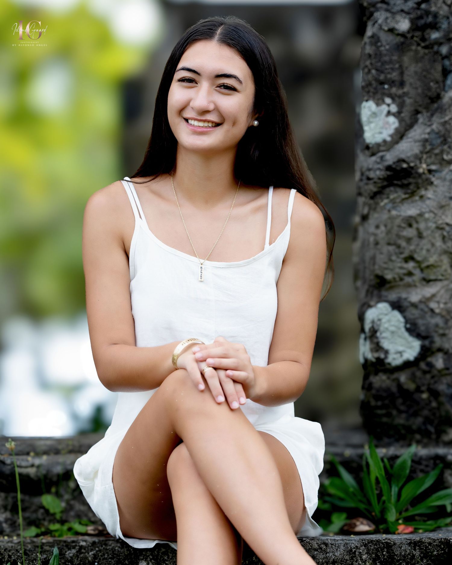A person in a white summer dress sits outdoors against a stone wall with lush greenery in the background.