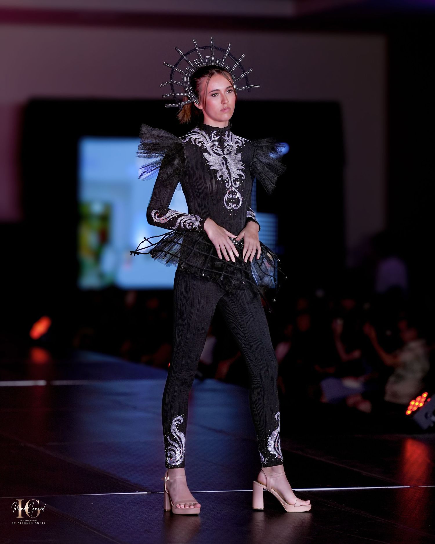 Avant-garde black outfit with ornate headpiece and silver embellishments presented on fashion runway.