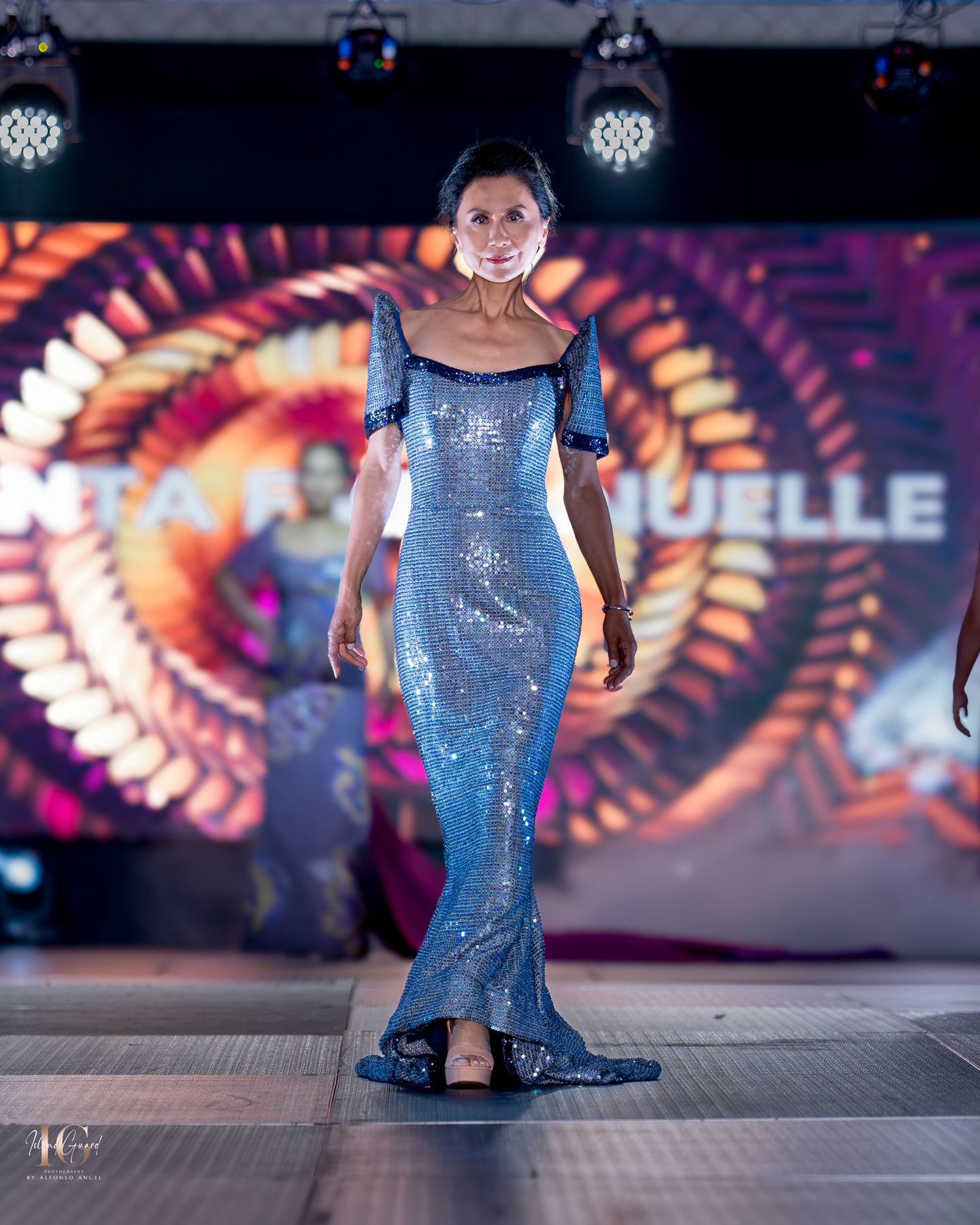 Sparkling blue mermaid gown with off-shoulder neckline presented on illuminated runway stage.