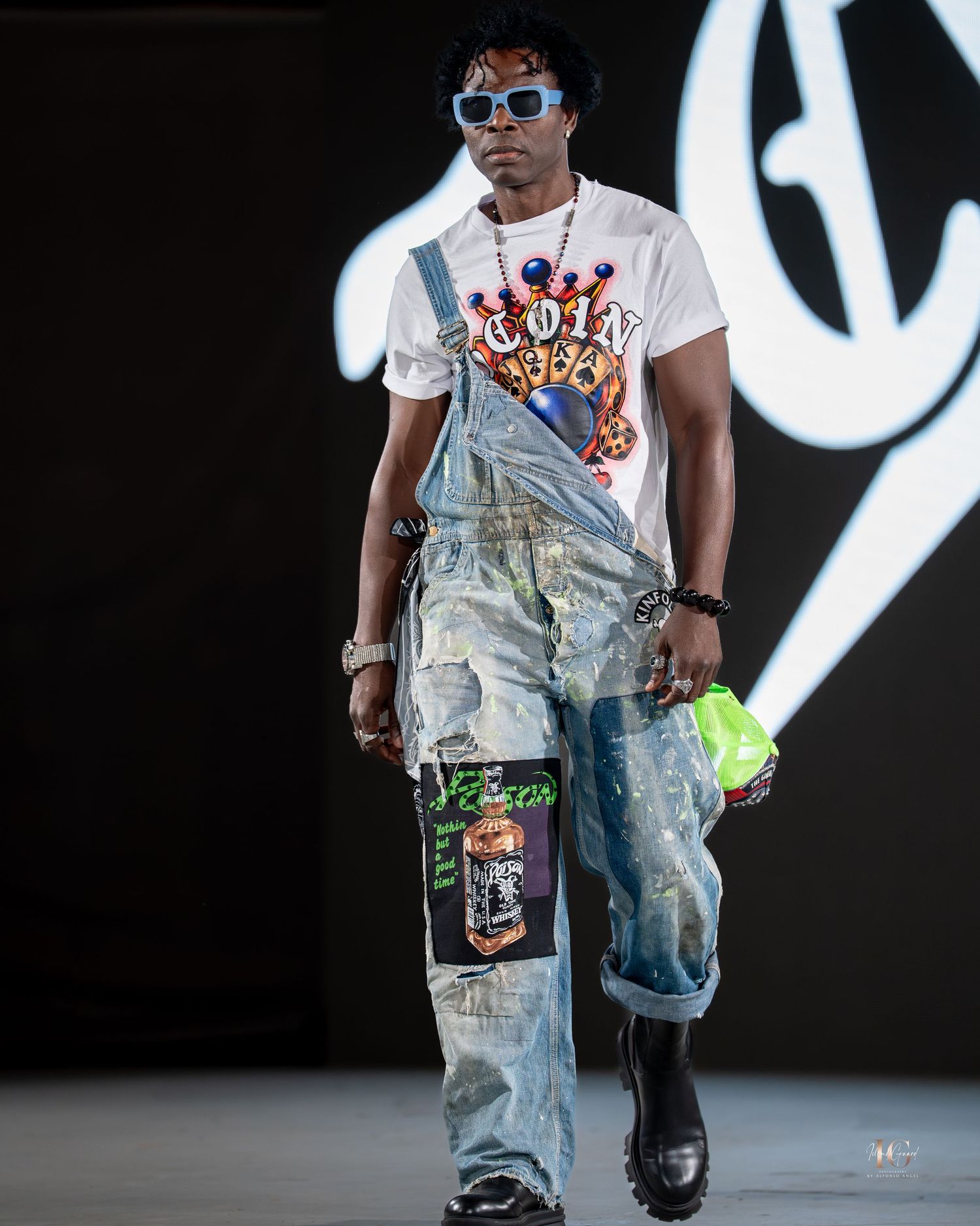 Model walks runway in distressed denim overalls and white t-shirt during fashion show against dark background.
