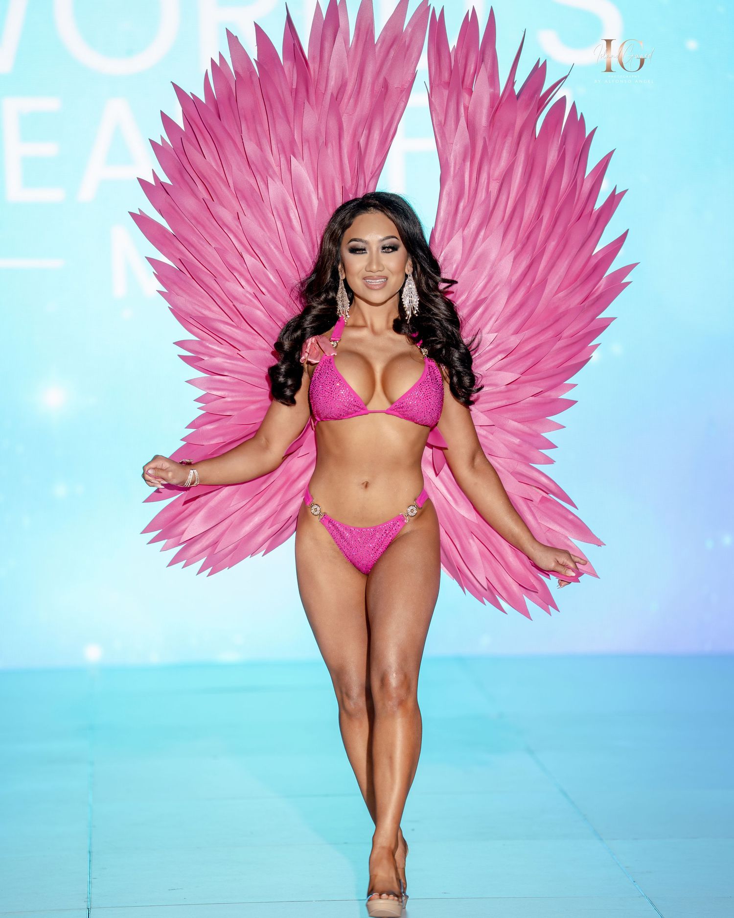 Model walks runway in bright pink bikini with large feathered angel wing props at fashion show.