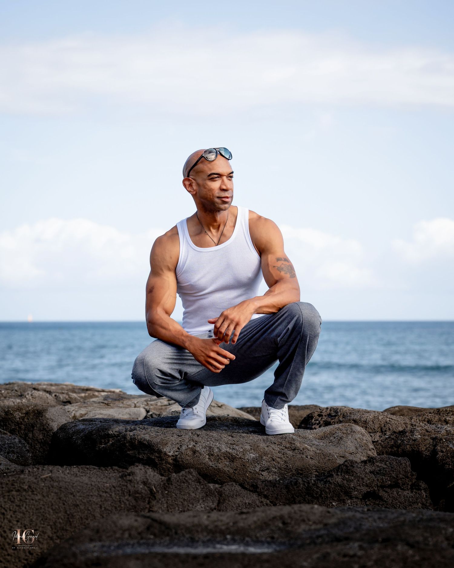 Athletic man in white tank top sits on black volcanic rock formations by the ocean on a bright sunny day.