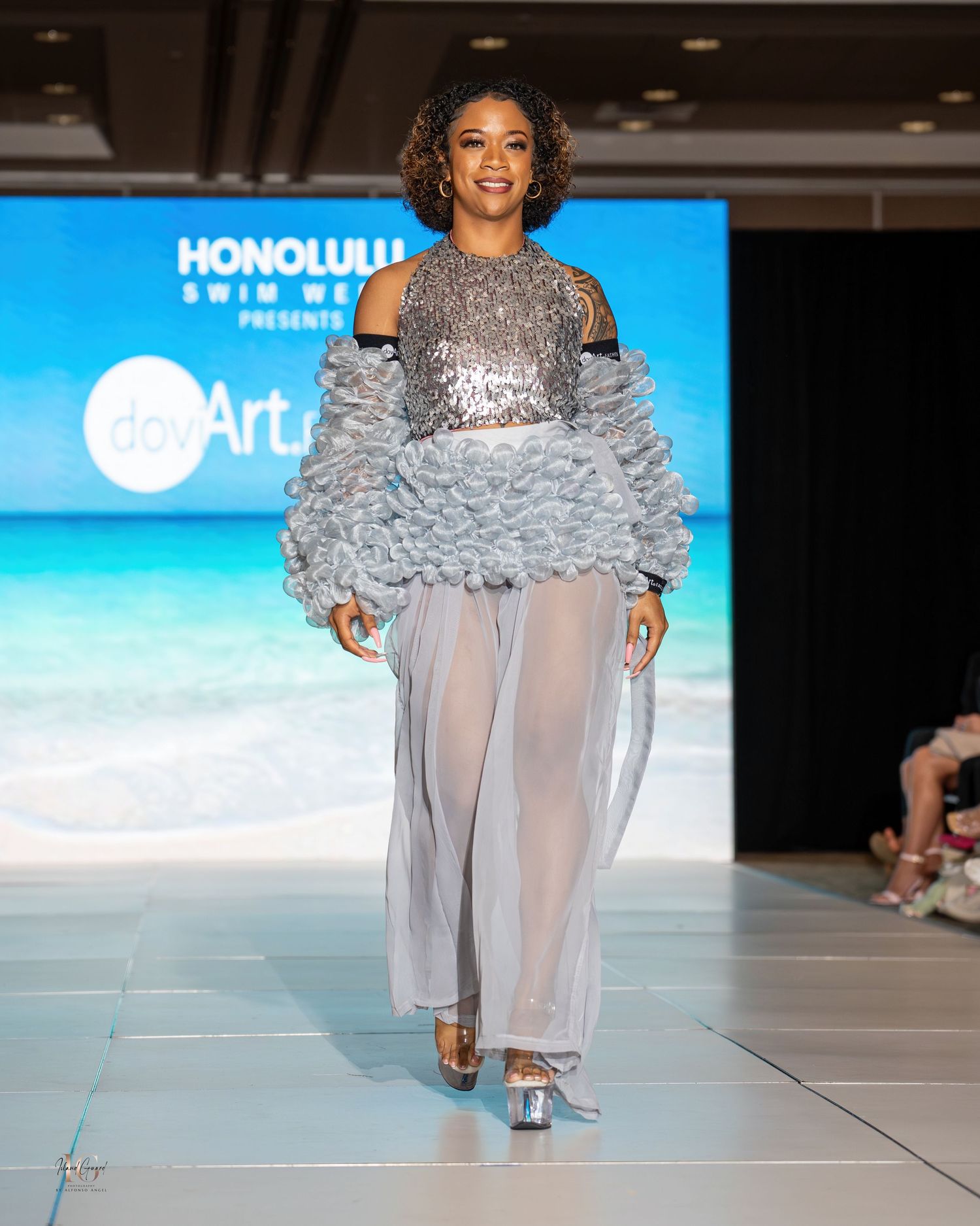 Fashion model presents silver sequined top with gray ruffled sleeves and sheer pants on catwalk.