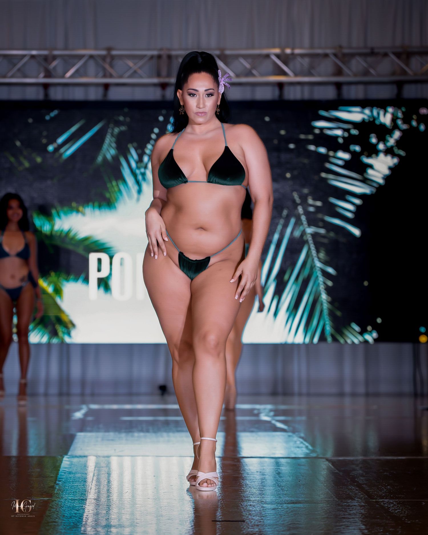 Curvy model presents black swimwear collection against tropical backdrop on runway.