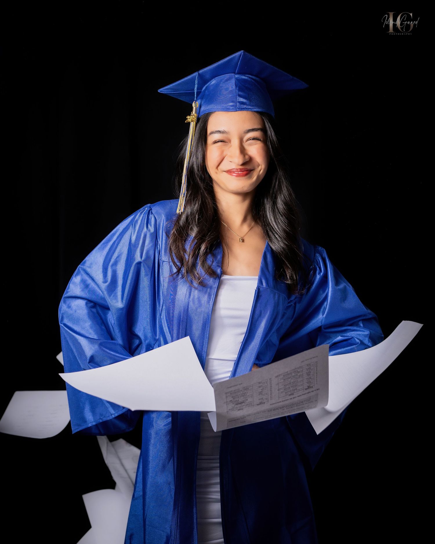 A graduate in blue cap and gown holding papers photographed against a black background.