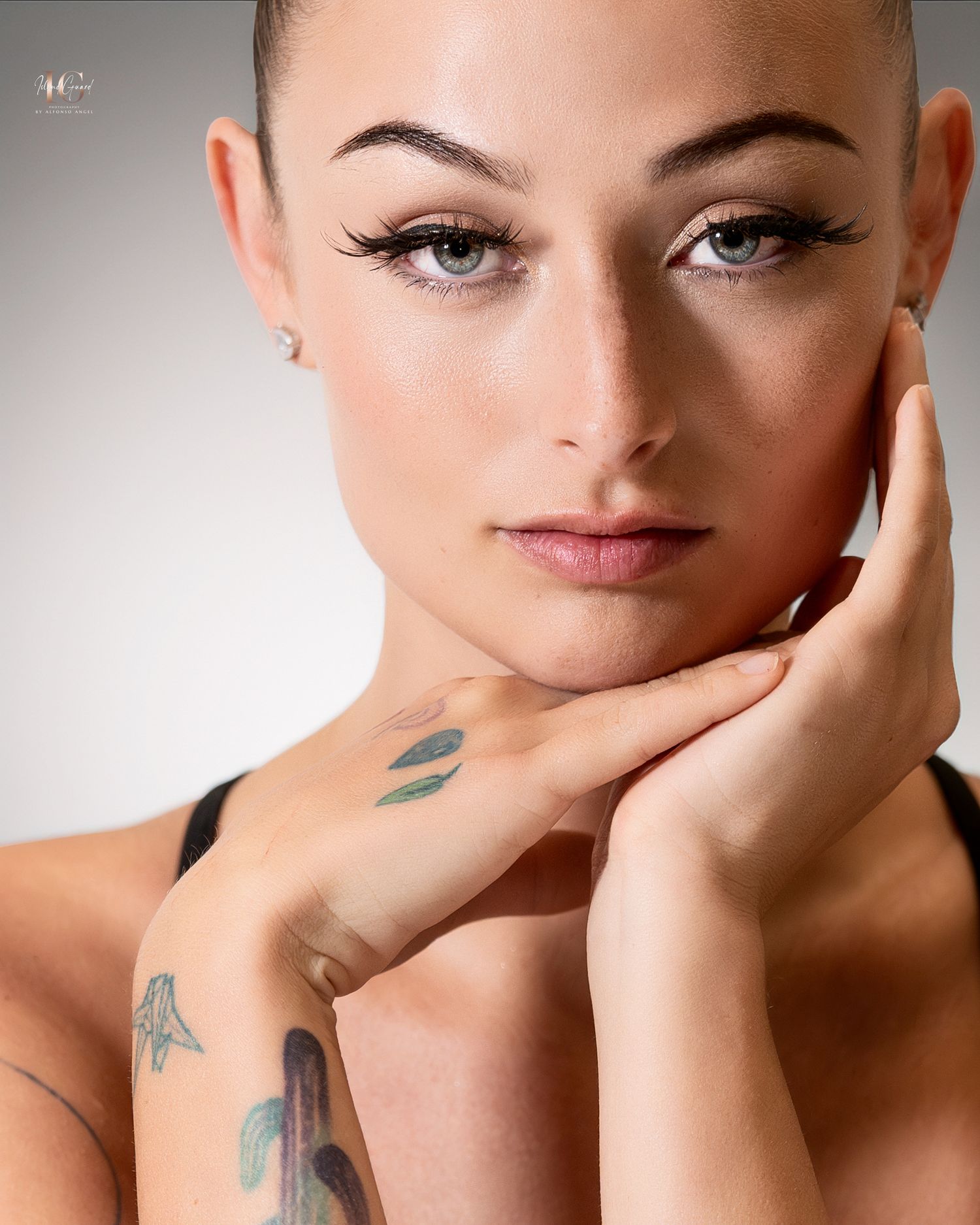 Portrait with dramatic winged eyeliner, soft neutral makeup, and visible tattoos on hand and neck.
