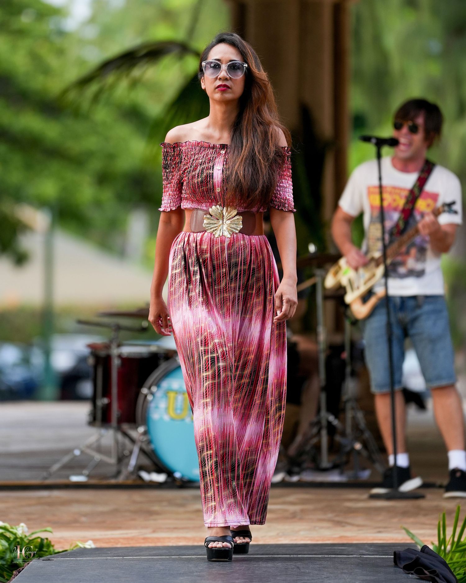 A person in a pink tie-dye dress and sunglasses walks along a tree-lined path during an outdoor event.