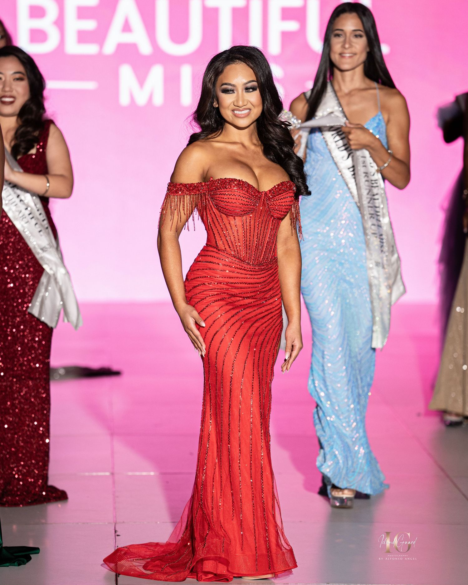 A model walks the runway in a flowing red evening gown during a fashion show against a bright pink backdrop.