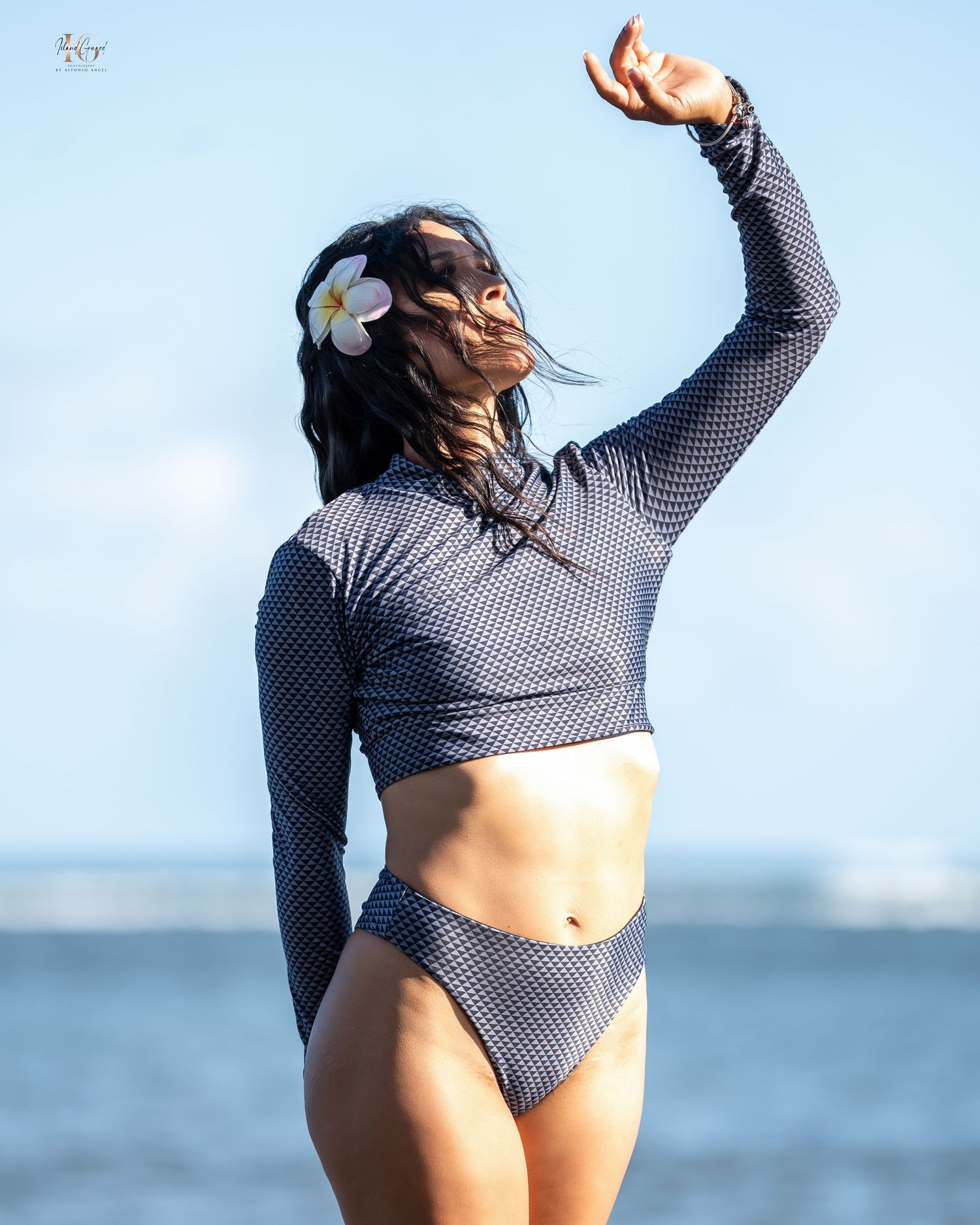 Athletic figure in a long sleeve mesh crop top and matching bottoms poses gracefully by the ocean with a plumeria flower.