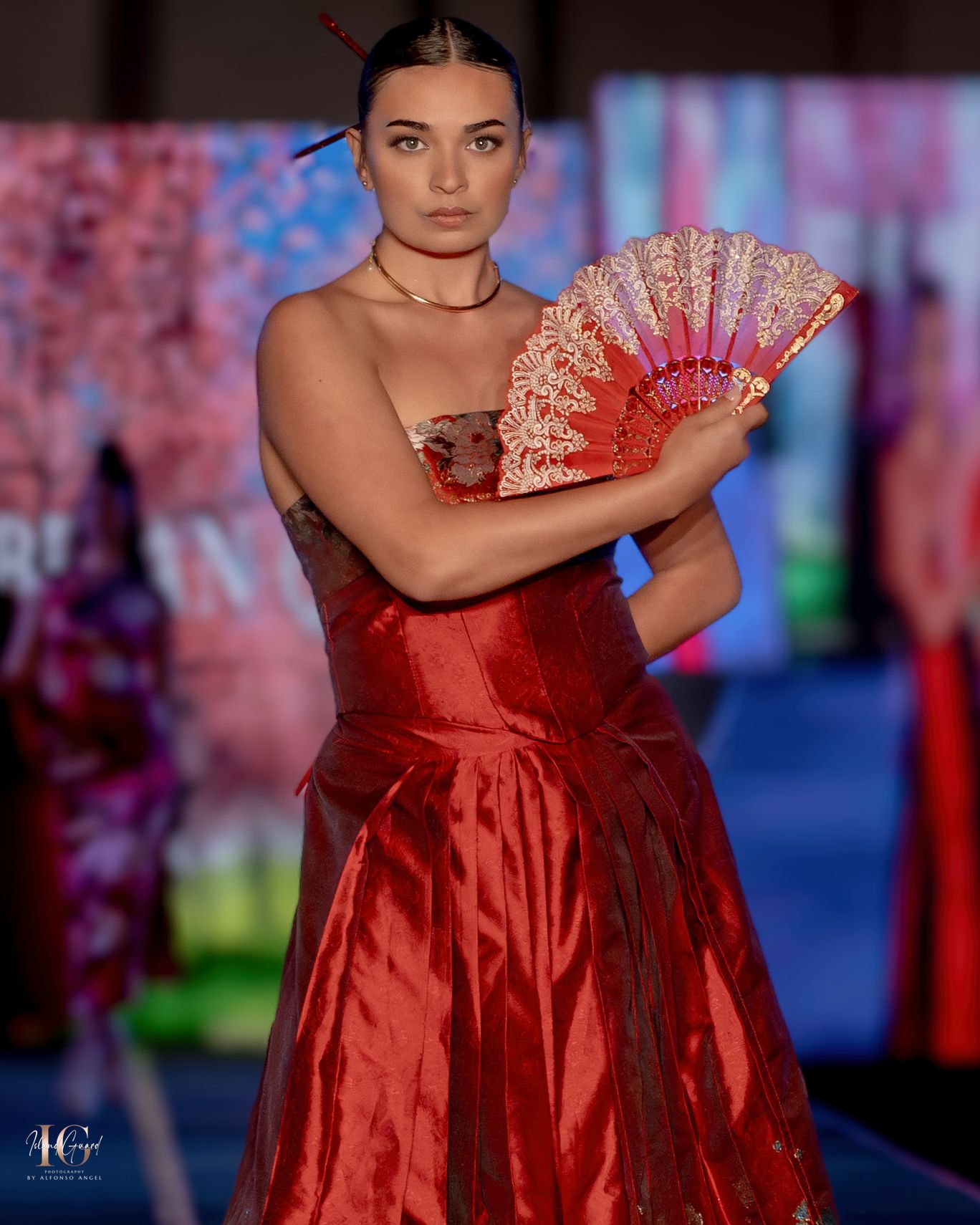 Model presents flowing red satin evening gown with matching fan on fashion runway.