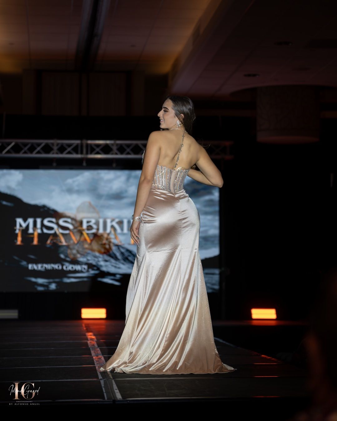 Model posing elegantly in a flowing champagne gown against dramatic lighting on stage during a pageant show.